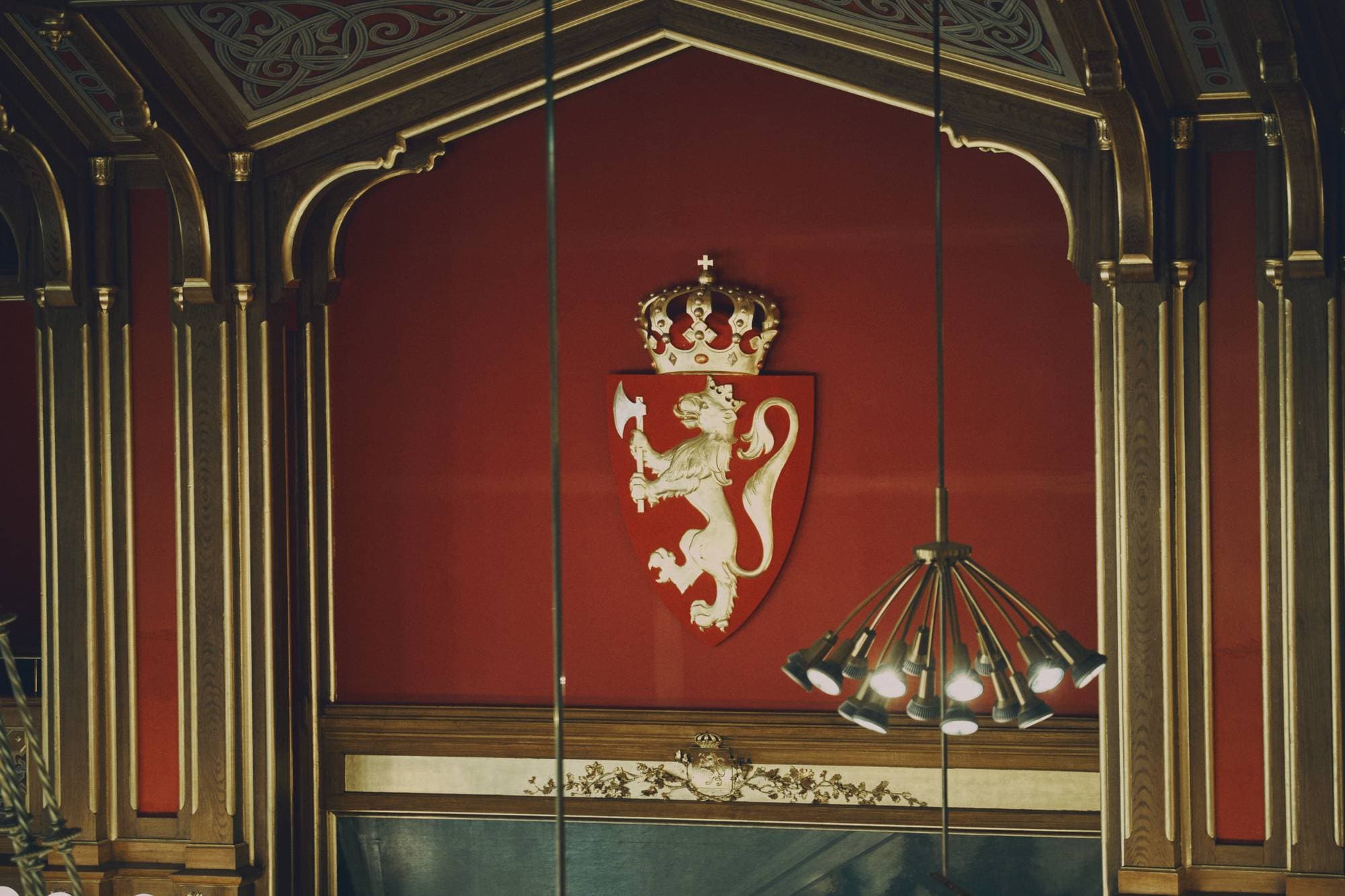 Inne på Stortinget. Rød vegg, og en lampe som henger fra veggen. Utsmykninger på veggene.