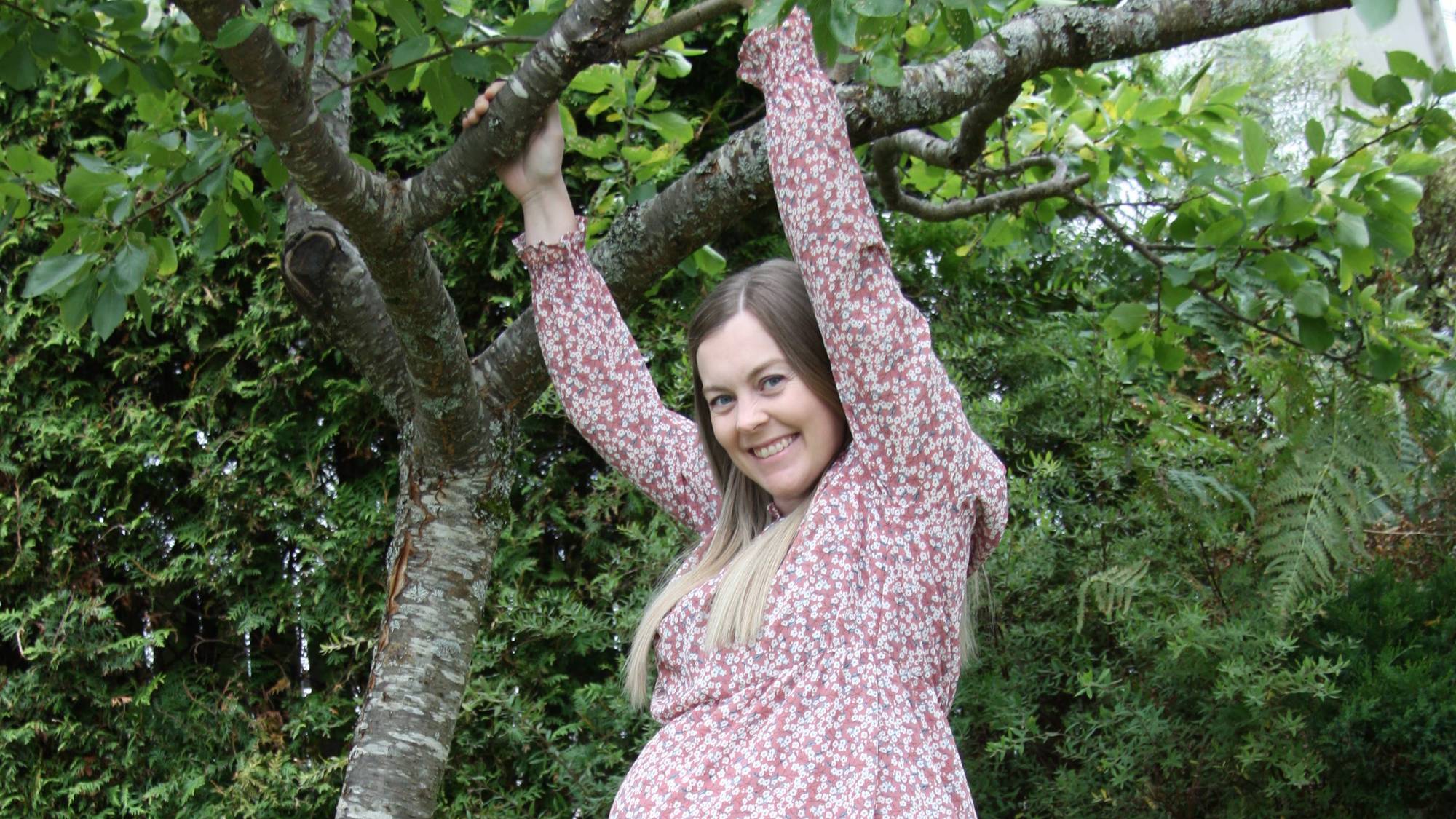 Gravid kvinne står ute i en hage i sommerkjole, med hendene i et tre og smiler til kamera.