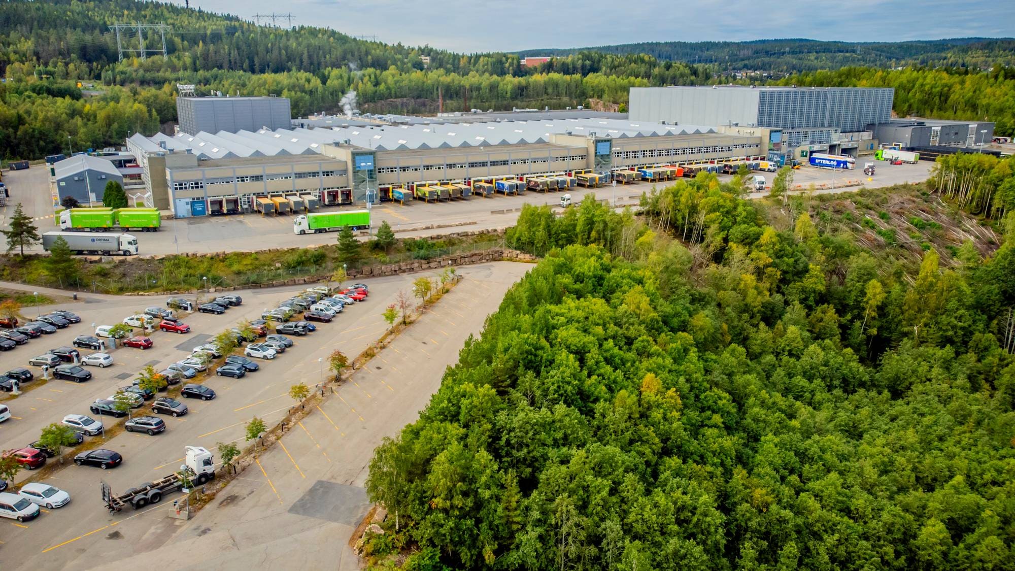 Flyfoto av stort industrianlegg med parkeringsplass og lastebiler, omgitt av skog. (Bildebeskrivelsen er laget av en KI-tjeneste)