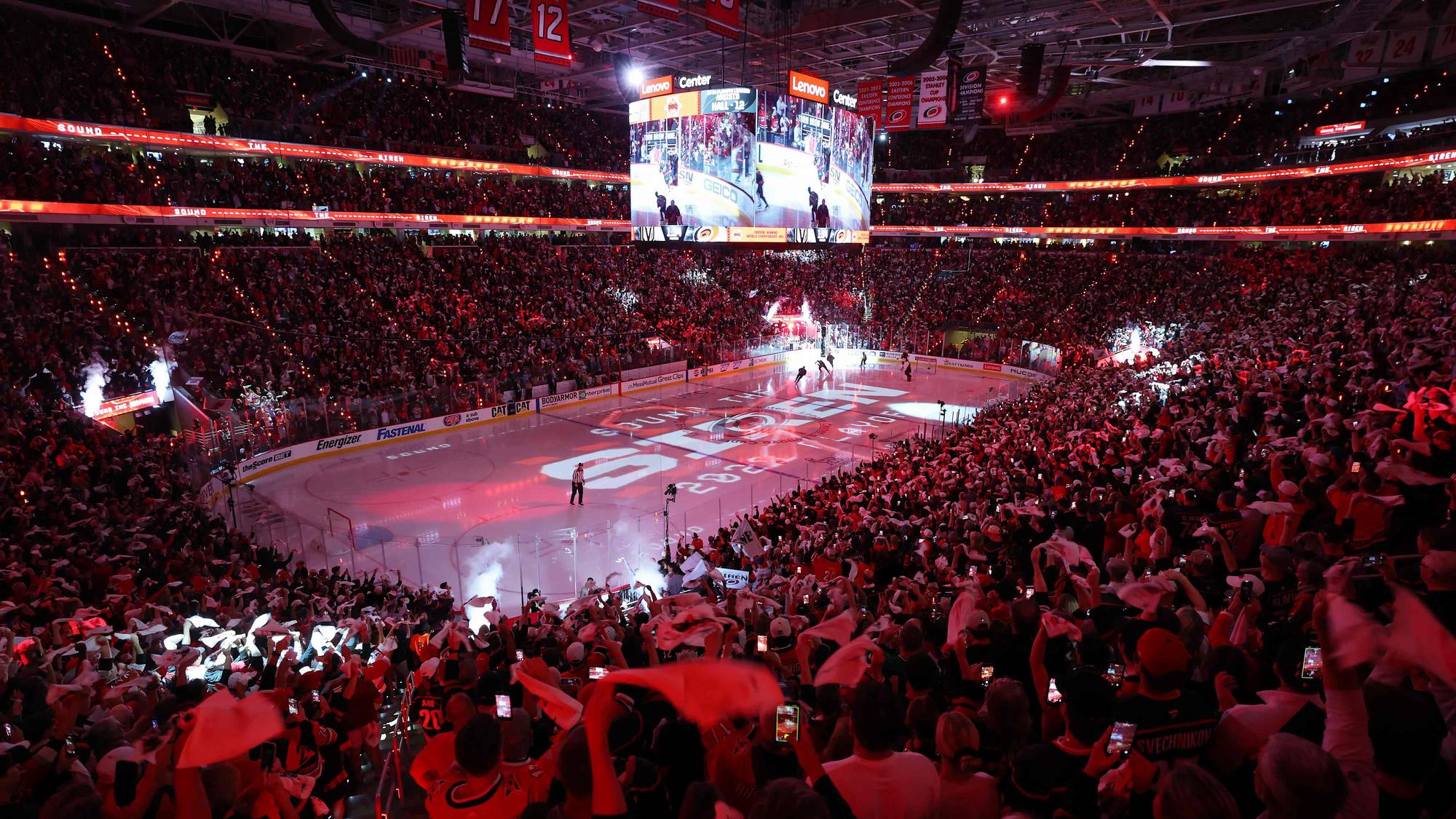 Publikum feirer i en fullsatt ishockeyarena mens spillere er på isen. Et stort videolerret viser action, og tilskuere holder opp lys og flagg. Rødt lys og konfetti fyller rommet. (Bildebeskrivelsen er laget av en KI-tjeneste)