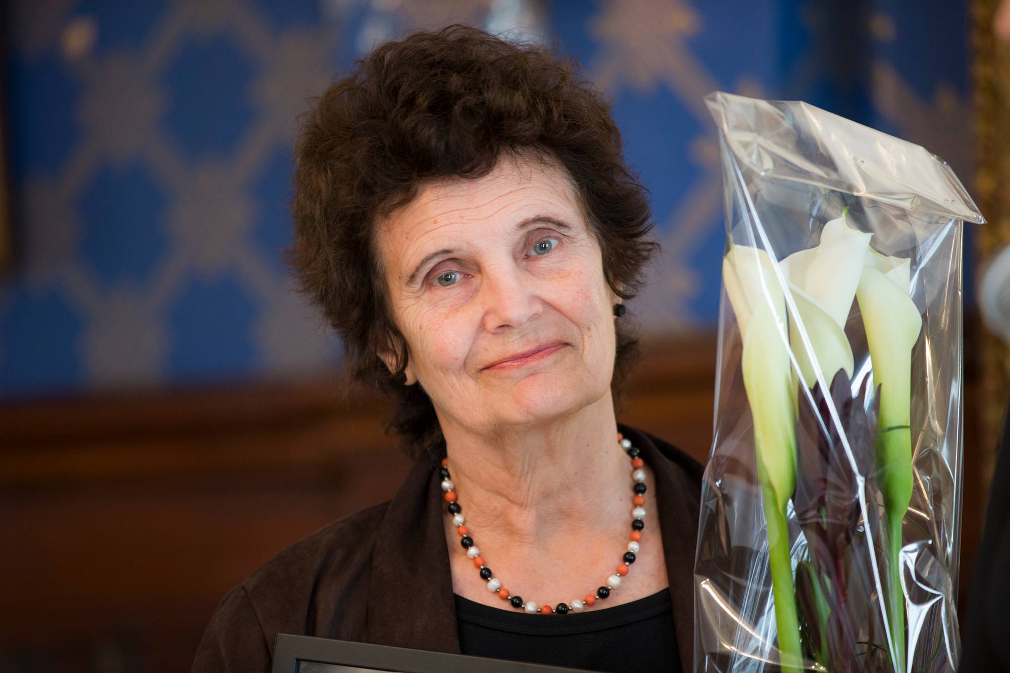 En kvinne med brunt krøllete hår og et perlekjede rundt halsen smiler. Hun holder en bukett med hvite blomster i plast ved siden av seg. Bak henne er en blå vegg med et mønster. (Bildebeskrivelsen er laget av en KI-tjeneste)
