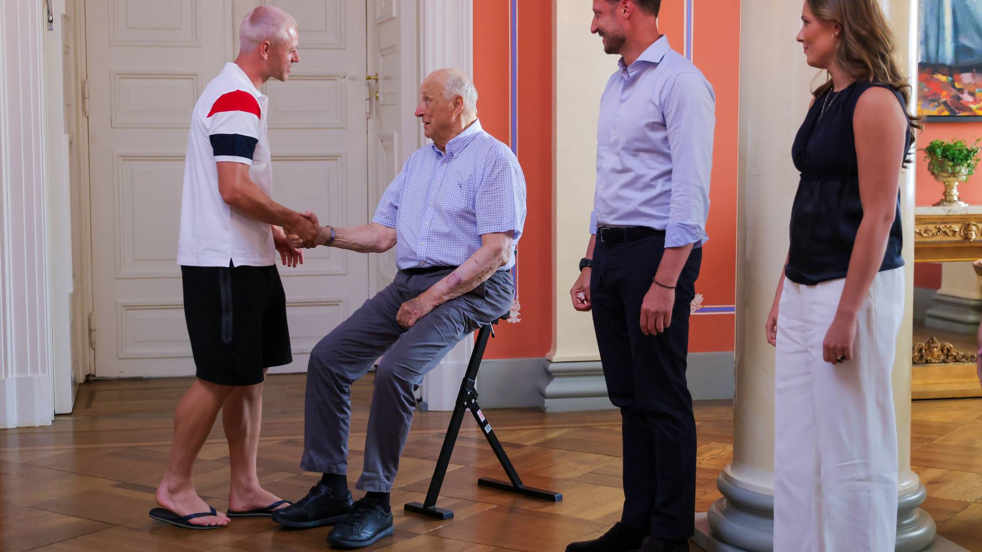 Mann i t-skjorte, shorts og flipflops hilser på kong Harald, Haakon og Ingrid-Alexandra