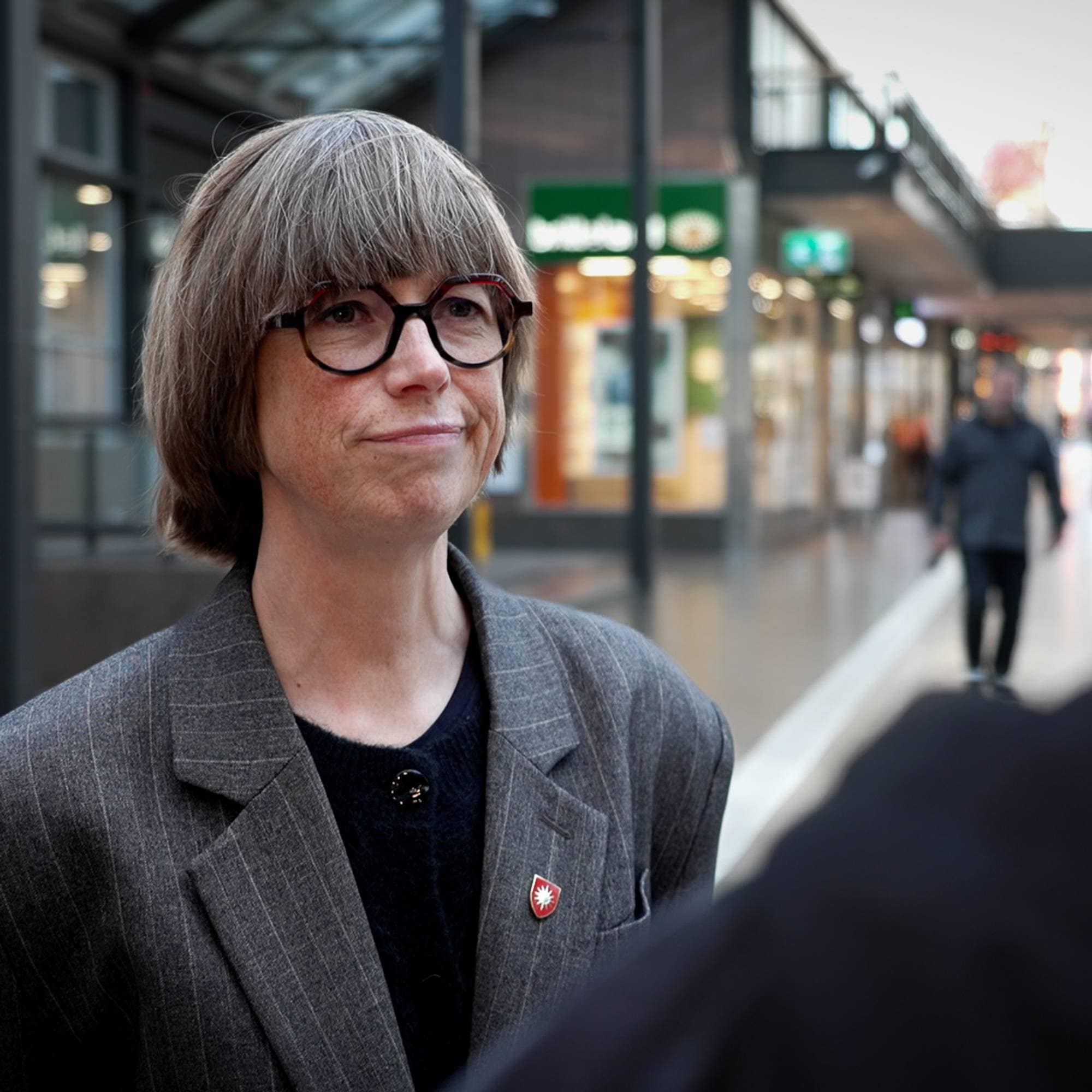 Kvinne med briller i samtale med en person ut av fokus i en handlegate. (Bildebeskrivelsen er laget av en KI-tjeneste)