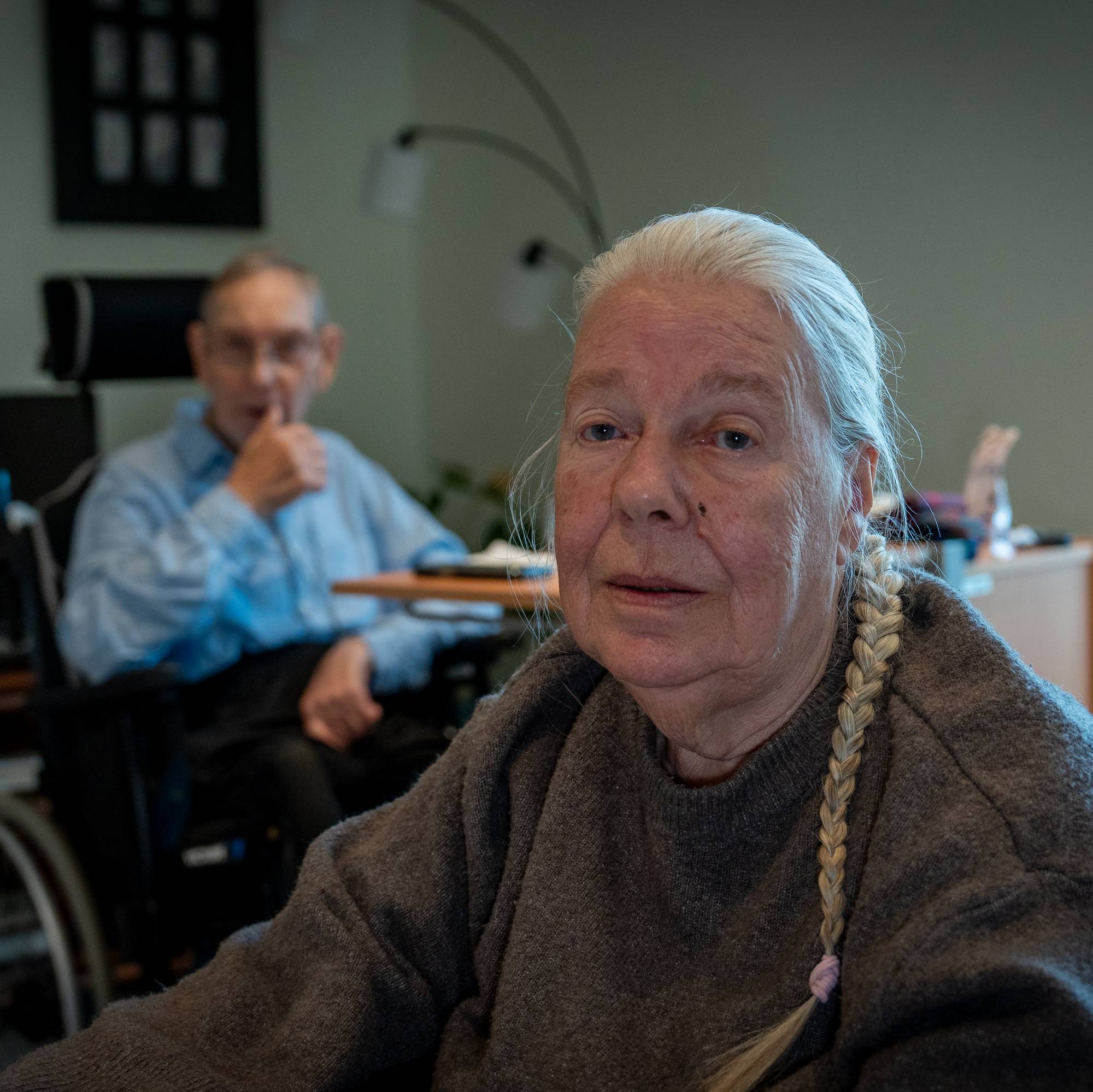 To eldre personer sitter i rullestoler i en stue. Den ene sitter i forgrunnen, den andre bak. (Bildebeskrivelsen er laget av en KI-tjeneste)