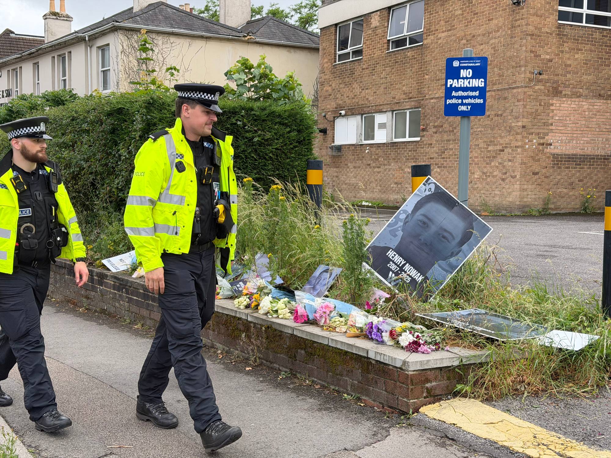 To politimenn i gule refleksveste går forbi en minnesmerkeplass. Minnesmerket består av blomster og flere bilder av en ung mann. Blant bildene er et stort portrett av en mann ved navn Henry Nowak. Bak minnesmerket er en mursteinsbygning med et "ingen parkering"-skilt. (Bildebeskrivelsen er laget av en KI-tjeneste)