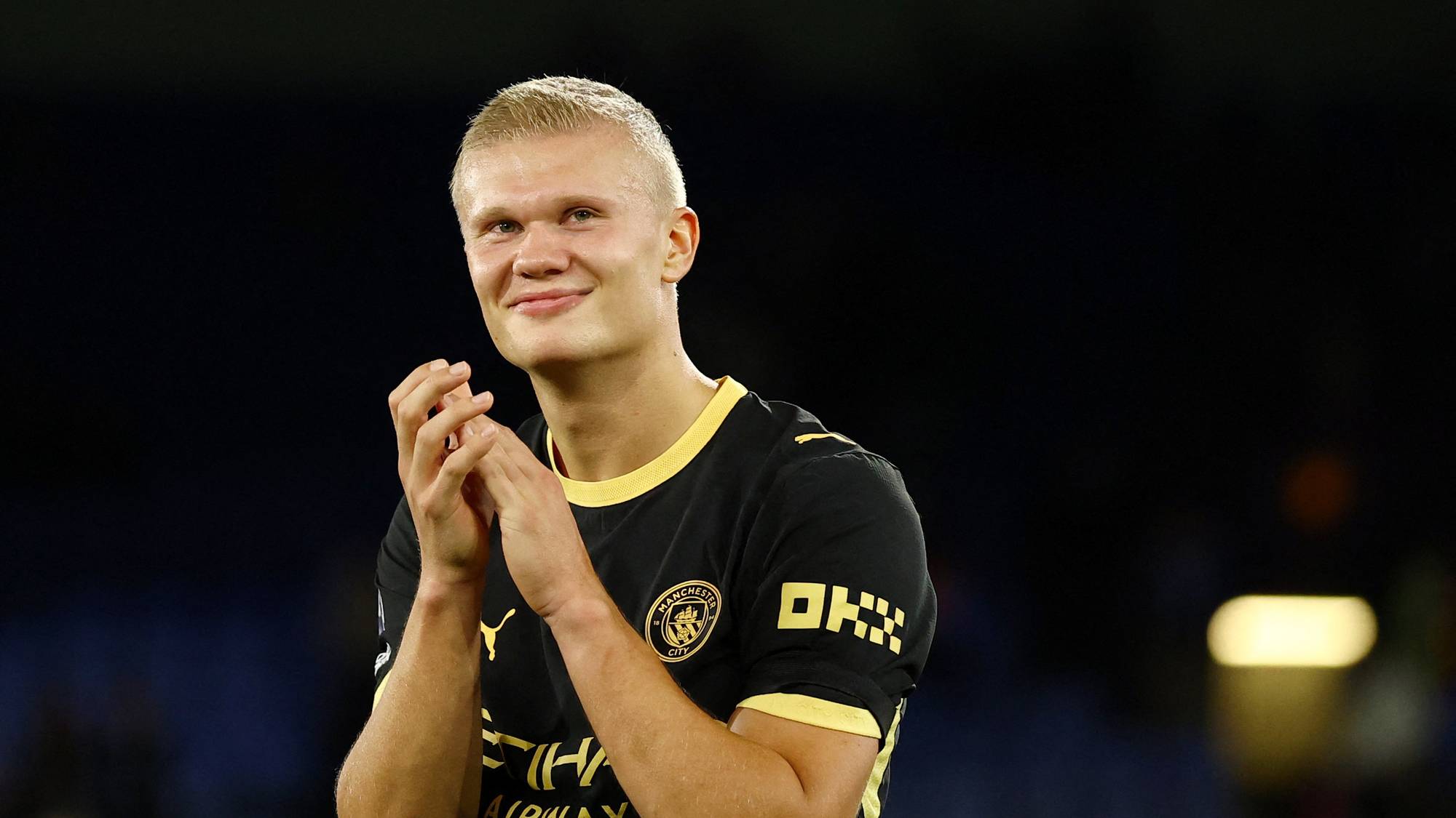Erling Braut Haaland i Manchester City-drakt klapper for publikum på en fotballstadion. (Bildebeskrivelsen er laget av en KI-tjeneste)
