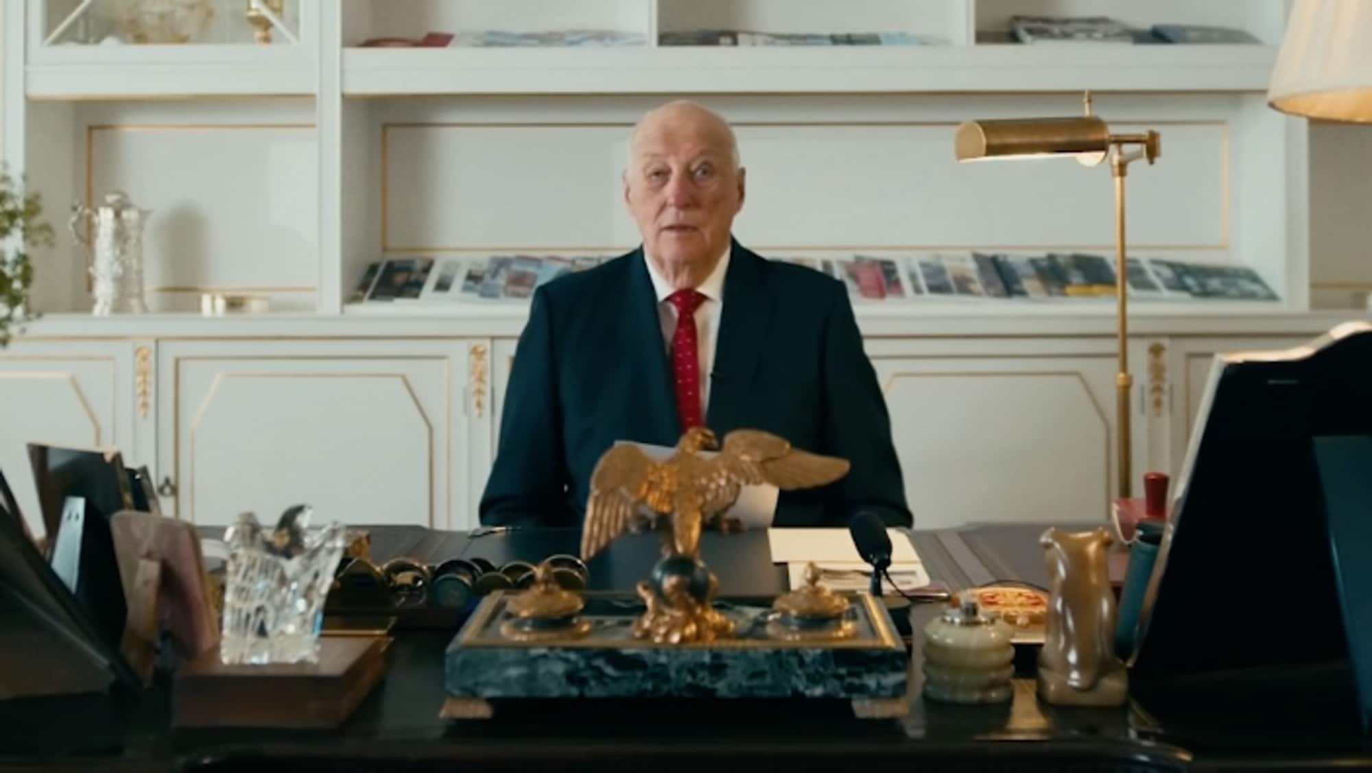 Kong Harald sitter bak en pult med flere pyntegjenstander