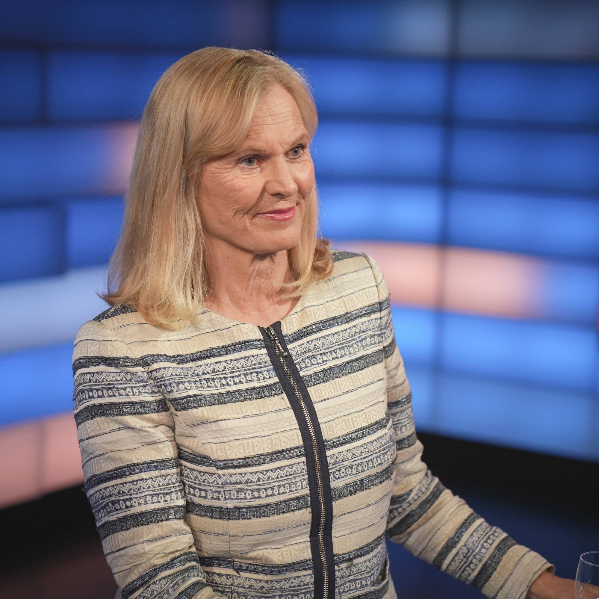 En blond kvinne i en mønstret jakke ved et bord i studio.