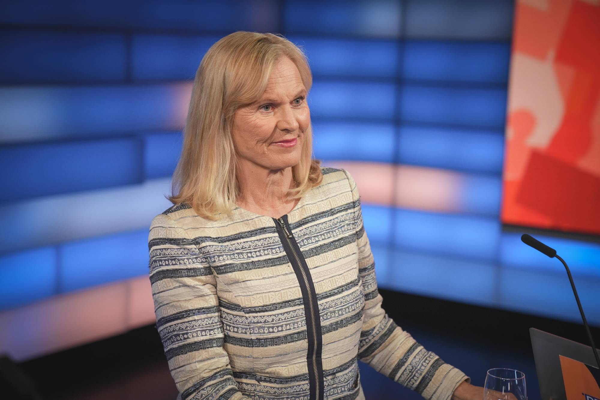 En blond kvinne i en mønstret jakke ved et bord i studio.
