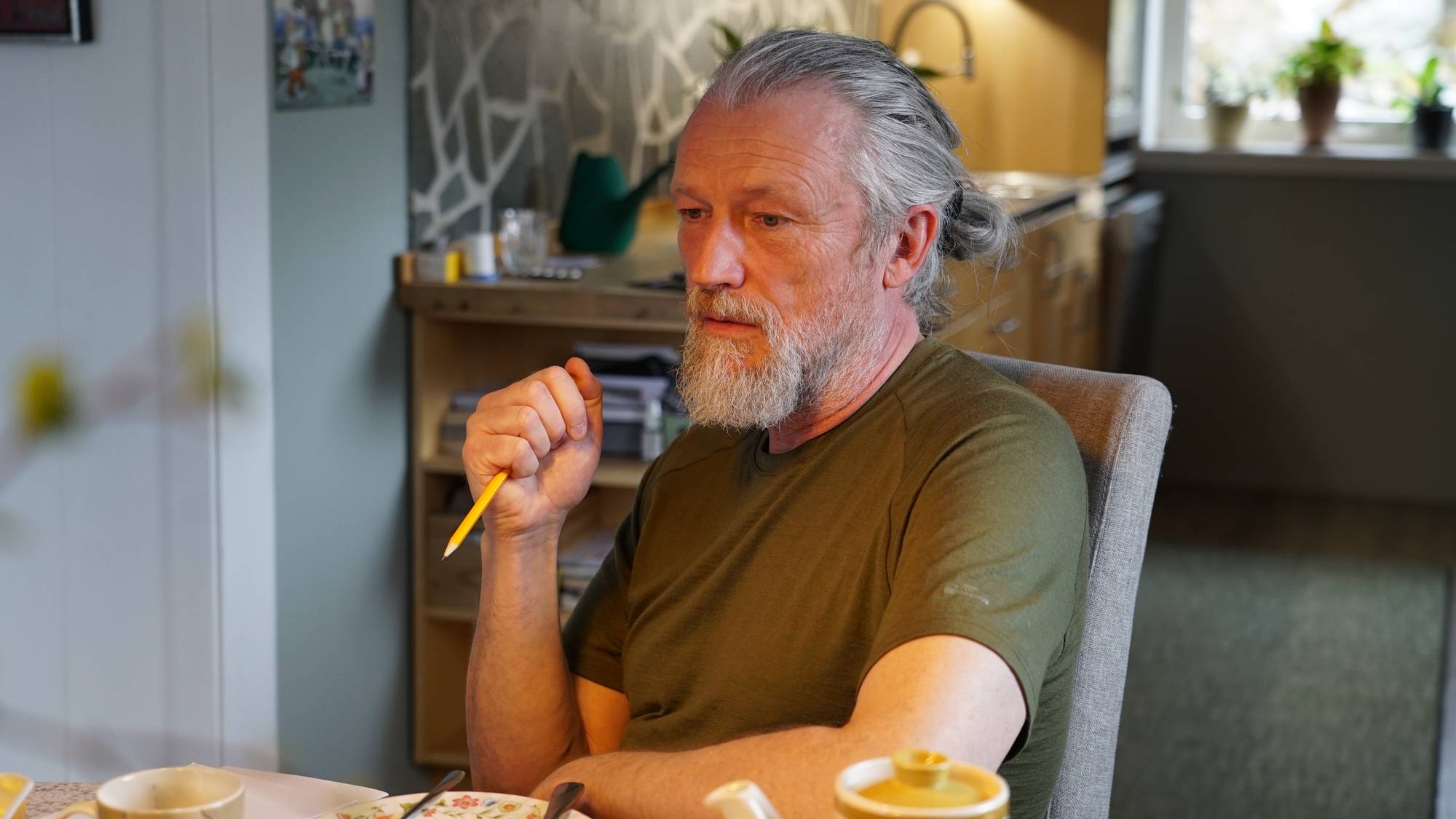 En mann med grått hår i hestehale og grønn t-skjorte som sitter ved et bord.
