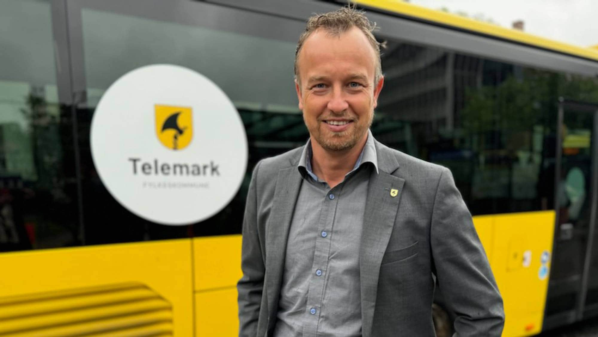 Vi ser en mann fra brystet og opp, posere foran en gul buss merket med Telemark. Han har brå dress over grå skjorte, kort litt bølgete hår, kort skjegg, og smiler. 