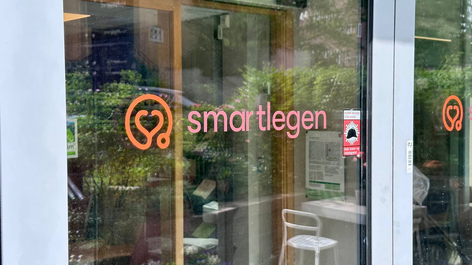 Tekst på glassdøren til en legetime er «smartlegen». Et rødt skilt med hjerte- og stetoskopmotiv og tekst «alarm tilknyttet 110 sentral» er synlig. Utenfor er grønt løvverk. (Bildebeskrivelsen er laget av en KI-tjeneste)