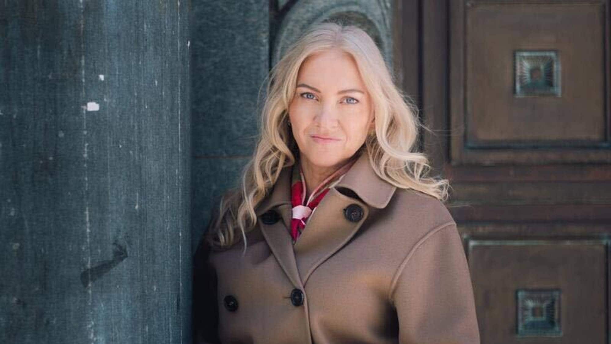 En blond middelaldrende kvinne ser i kamera. Hun har på seg en brun kåpe og står foran en inngangsdør.