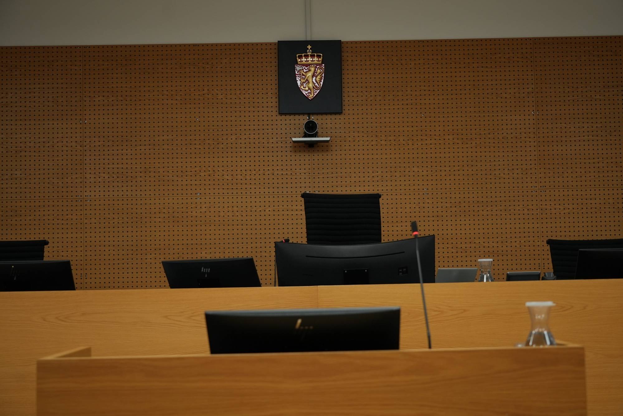 Tom rettssal med dommerplass, skjermer og riksvåpenet på veggen. (Bildebeskrivelsen er laget av en KI-tjeneste)
