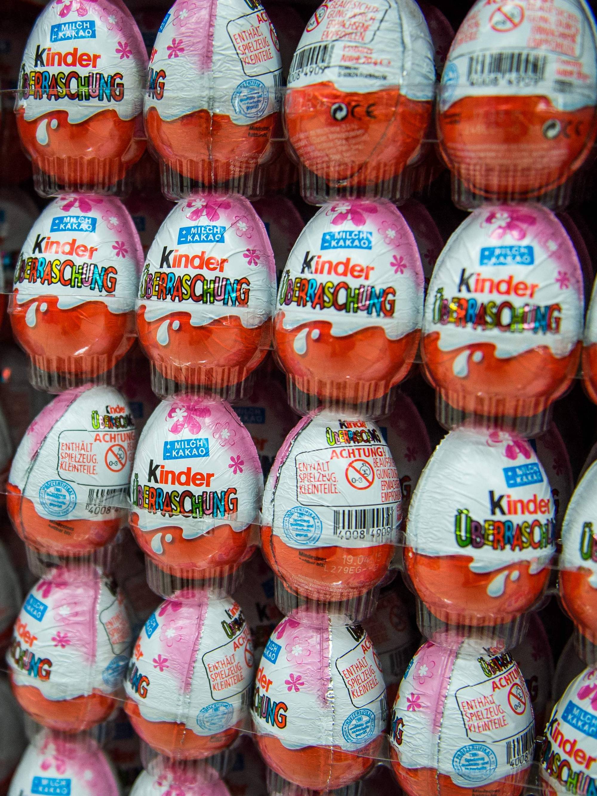 Bildet viser en stor mengde Kinder Überraschung-sjokolader stablet tett sammen. De er pakket i oransje og hvitt papir med tydelig merkevarelogo. Sjokoladene er presentert i et butikkvindu eller på en hylle. (Bildebeskrivelsen er laget av en KI-tjeneste)