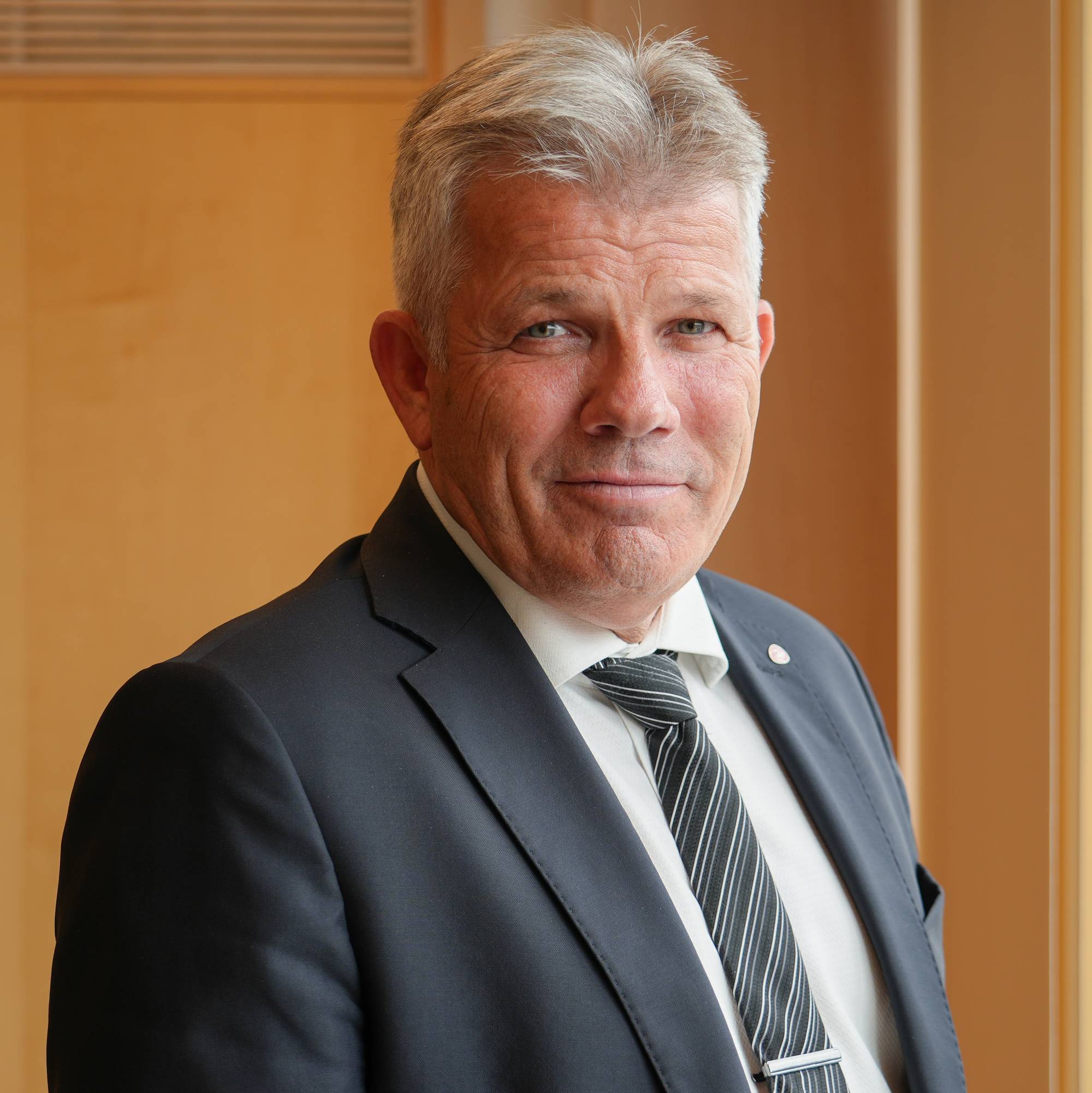 Mann med grått hår i dress og slips, stående ved et vindu. (Bildebeskrivelsen er laget av en KI-tjeneste)
