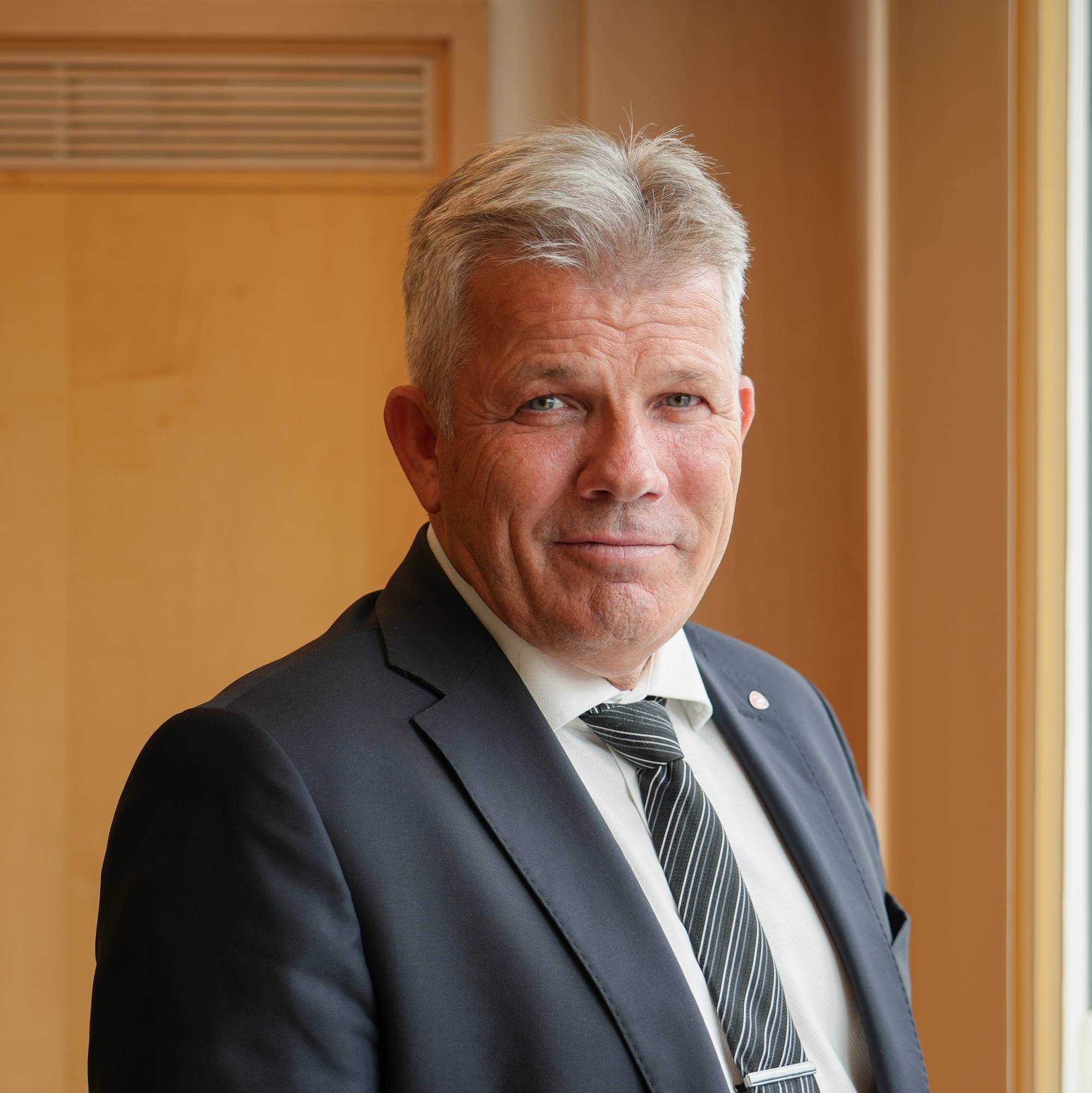 Mann med grått hår i dress og slips, stående ved et vindu. (Bildebeskrivelsen er laget av en KI-tjeneste)