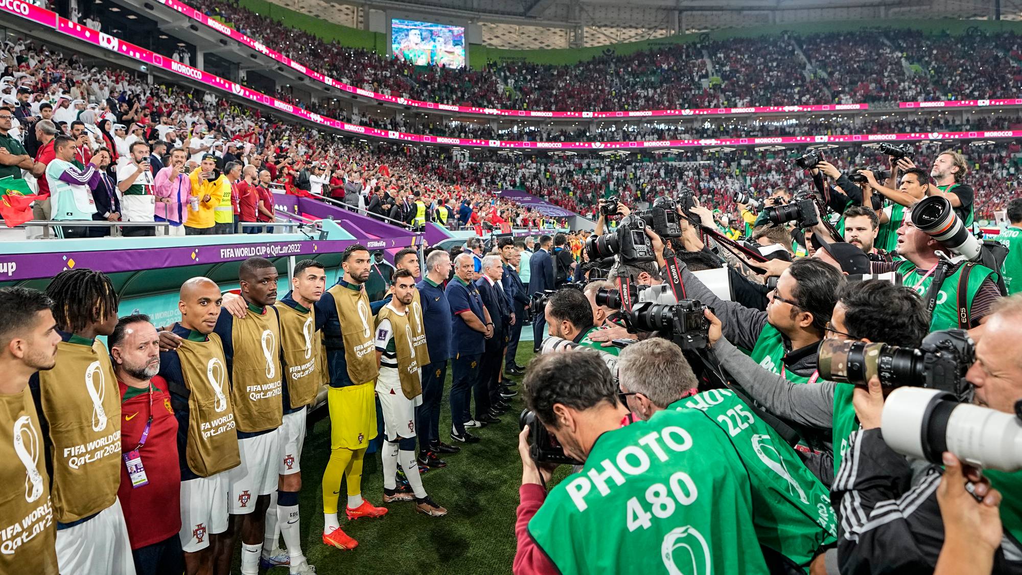 Fotballspillere står på rekke på en bane foran et publikum i en stadion. En gruppe fotografer med kameraer er samlet foran spillerne.
Publikum på tribunen er synlig bak spillerne og fotografene.
De fleste spillerne har på seg gule vest utenpå sine fotballdrakter.
En stor mengde mennesker dekker tribunene på stadion. (Bildebeskrivelsen er laget av en KI-tjeneste)
