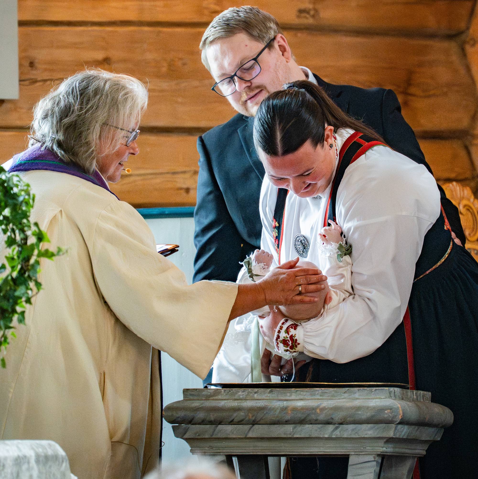 Prest døper baby i en kirke. Foreldre og faddere i bunad ser på under seremonien. (Bildebeskrivelsen er laget av en KI-tjeneste)