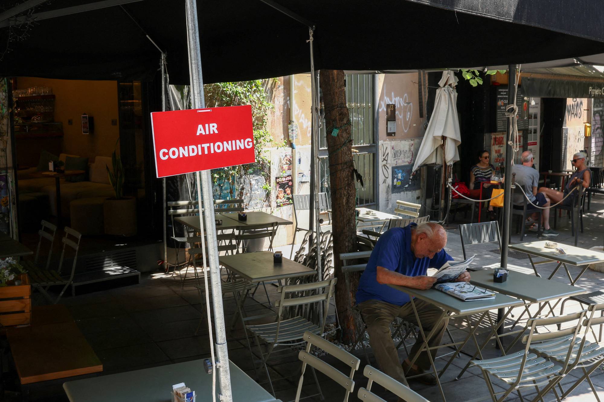 En mann sitter ved et kafébord utendørs under et skilt med teksten Air conditioning. (Bildebeskrivelsen er laget av en KI-tjeneste)