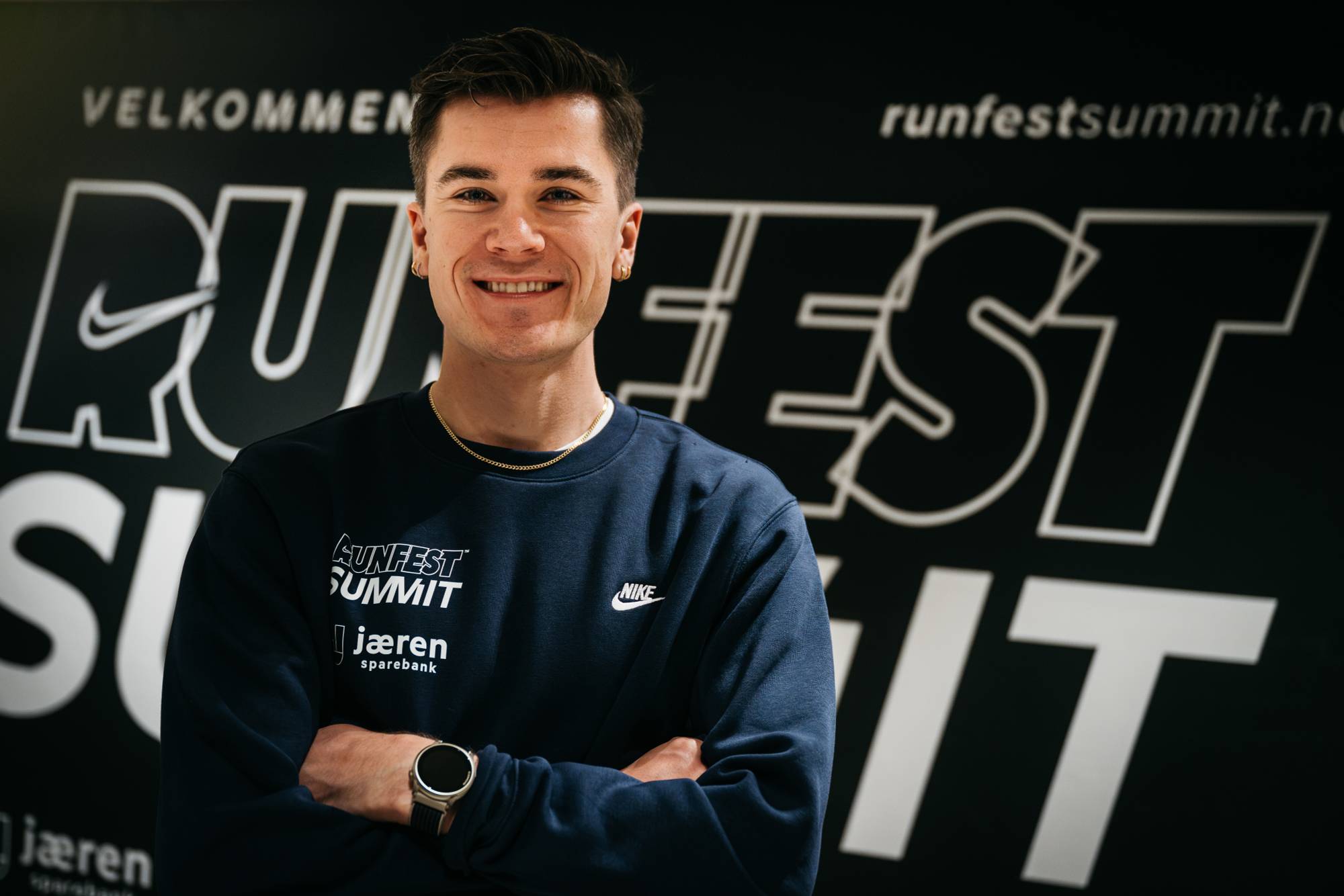 En smilende mann med armene i kors foran en Runfest Summit-vegg. (Bildebeskrivelsen er laget av en KI-tjeneste)