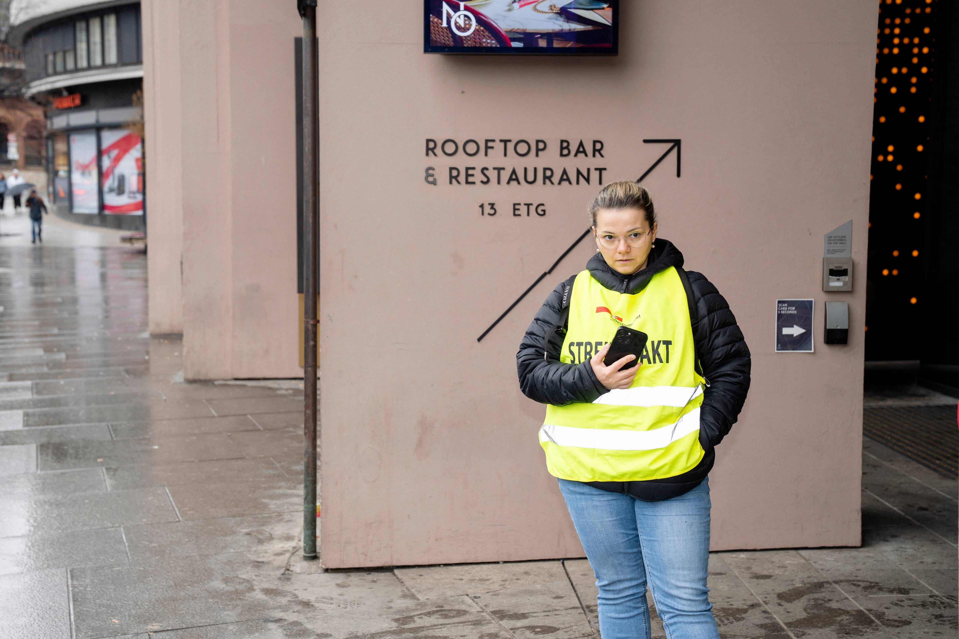 En kvinne med refleksvest og briller står utenfor en bygning. Hun holder en telefon i hånden. Bak henne er det skilt som viser "ROOFTOP BAR & RESTAURANT". Utenfor er det en våt gate med brostein og bygninger. (Bildebeskrivelsen er laget av en KI-tjeneste)