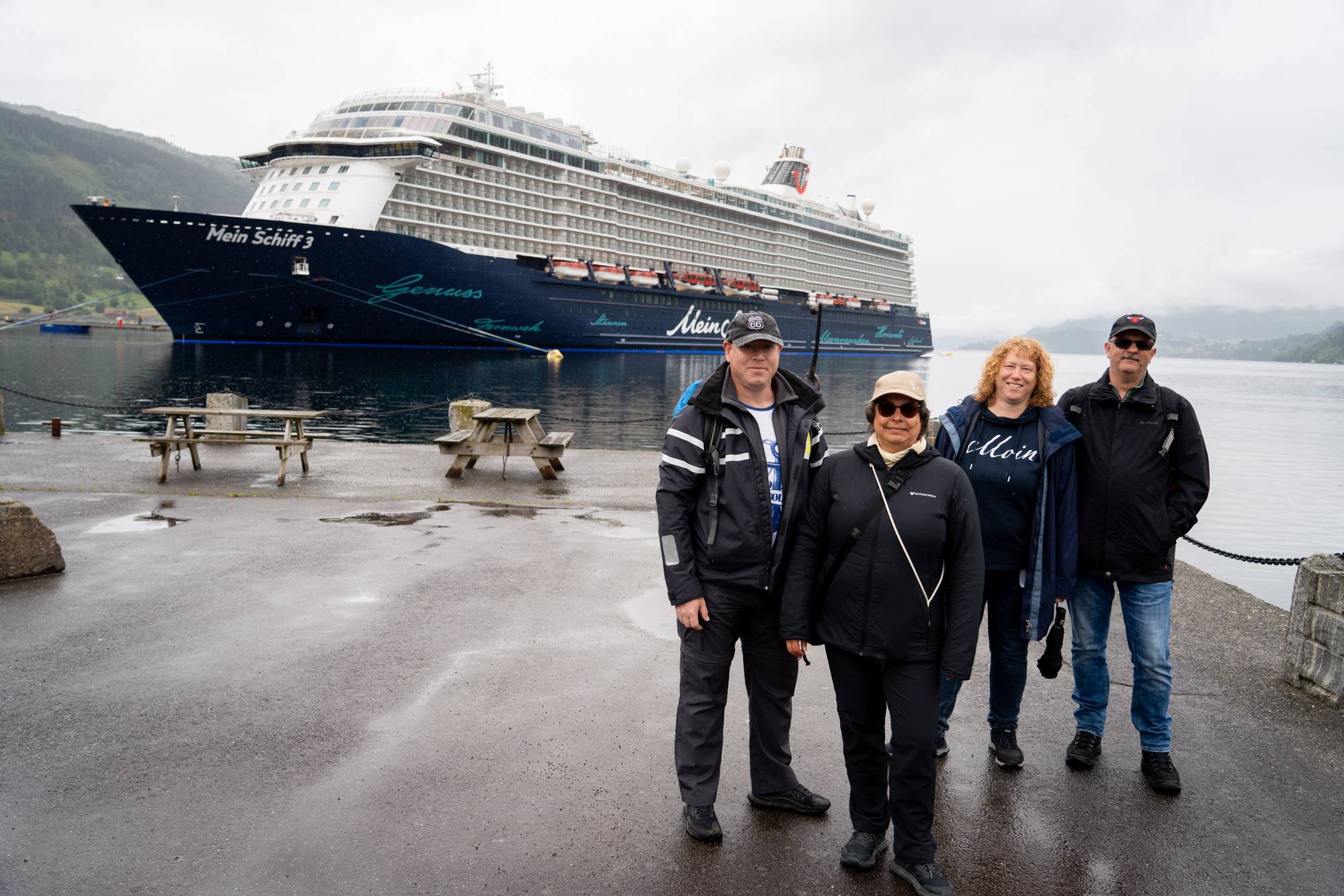 Fire personer står på kaien foran cruiseskipet Mein Schiff 3. Skipet ligger til kai i en fjord, med fjell i bakgrunnen. Været er overskyet. Det er benker og bord på kaia. (Bildebeskrivelsen er laget av en KI-tjeneste)