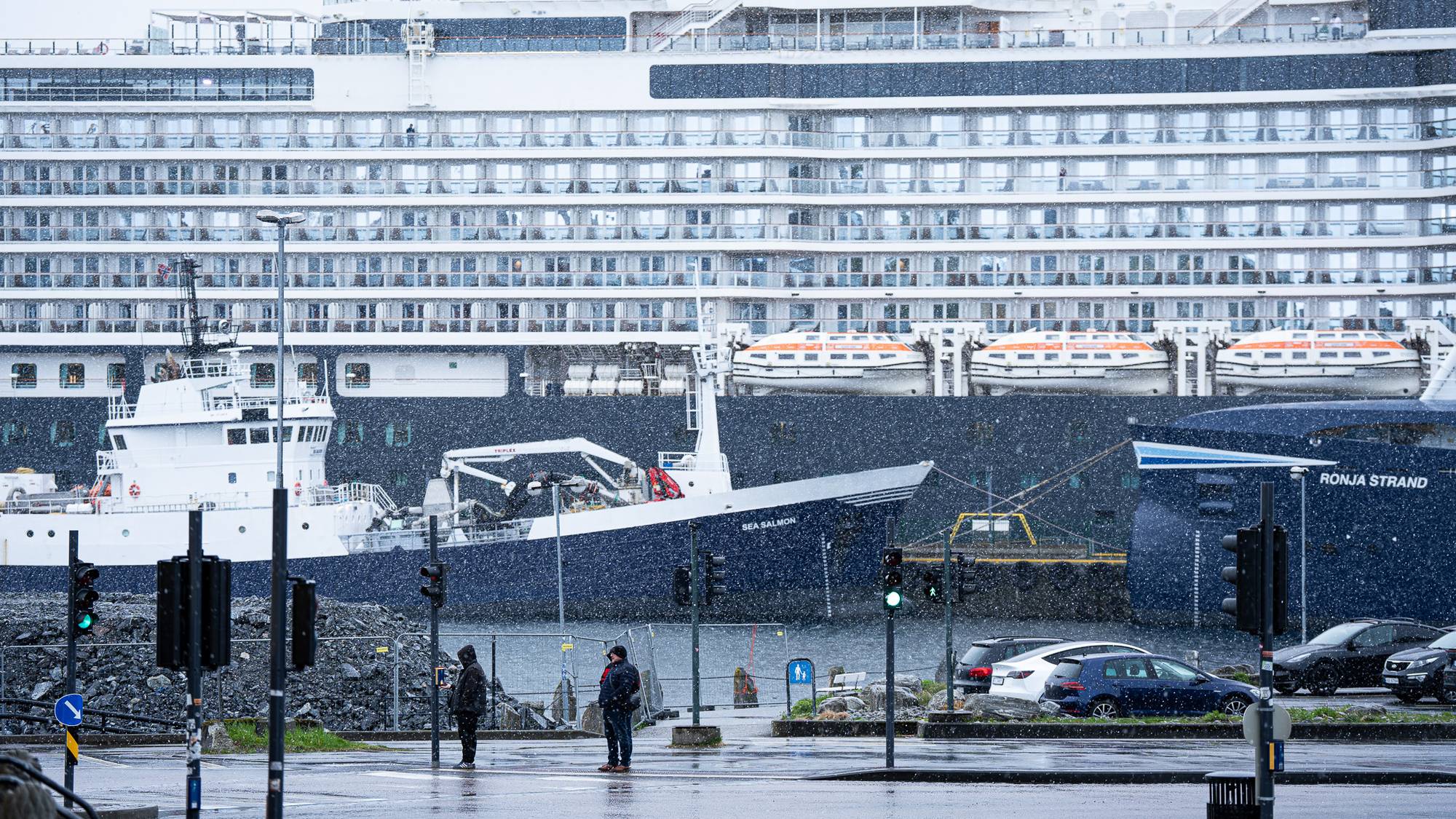 Et stort cruiseskip, Queen Anne, ligger ved kai. Foran cruiseskipet ligger et mindre skip ved navn Sea Salmon. Det snør, og to personer står ved et veikryss. (Bildebeskrivelsen er laget av en KI-tjeneste)