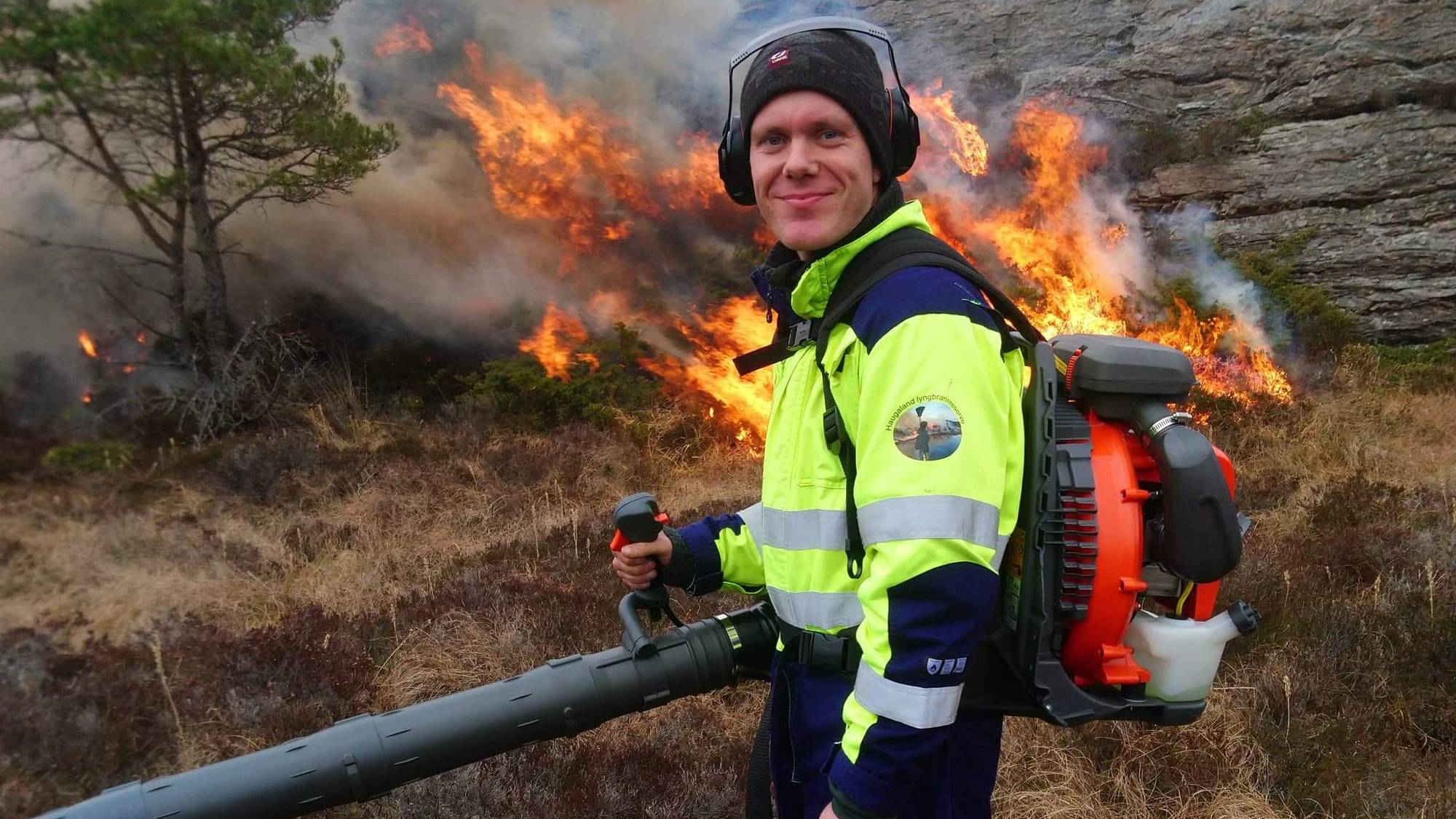 Ein mann i vernetøy står med ein lauvblåsar framfor ein kontrollert brenning i eit utmarksområde. 