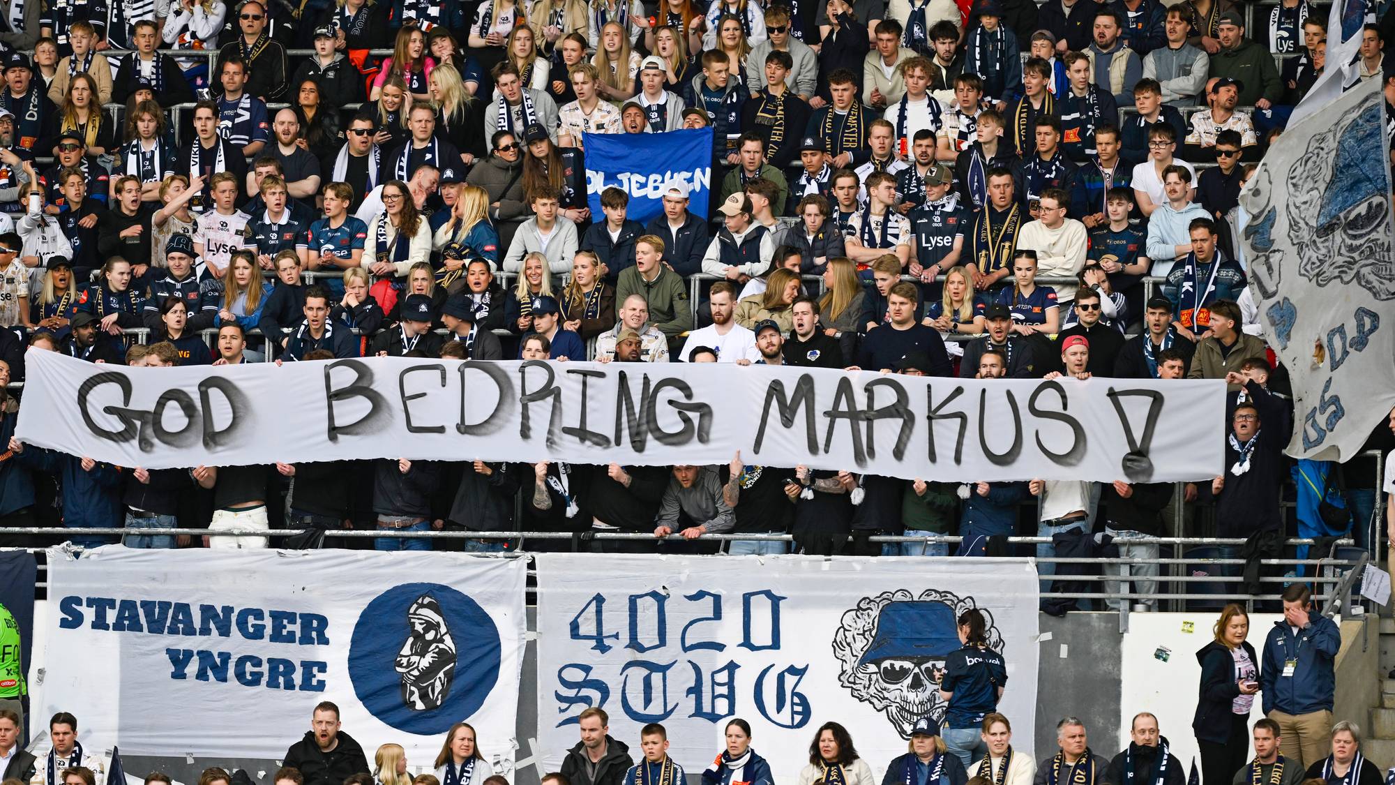 Tilskuere på en tribun av en stadion holder et banner med teksten "GOD BEDRING MARKUS". Flere personer i publikum har på seg supporterutstyr. Bak banneret er det flere andre bannere og flagg. (Bildebeskrivelsen er laget av en KI-tjeneste)