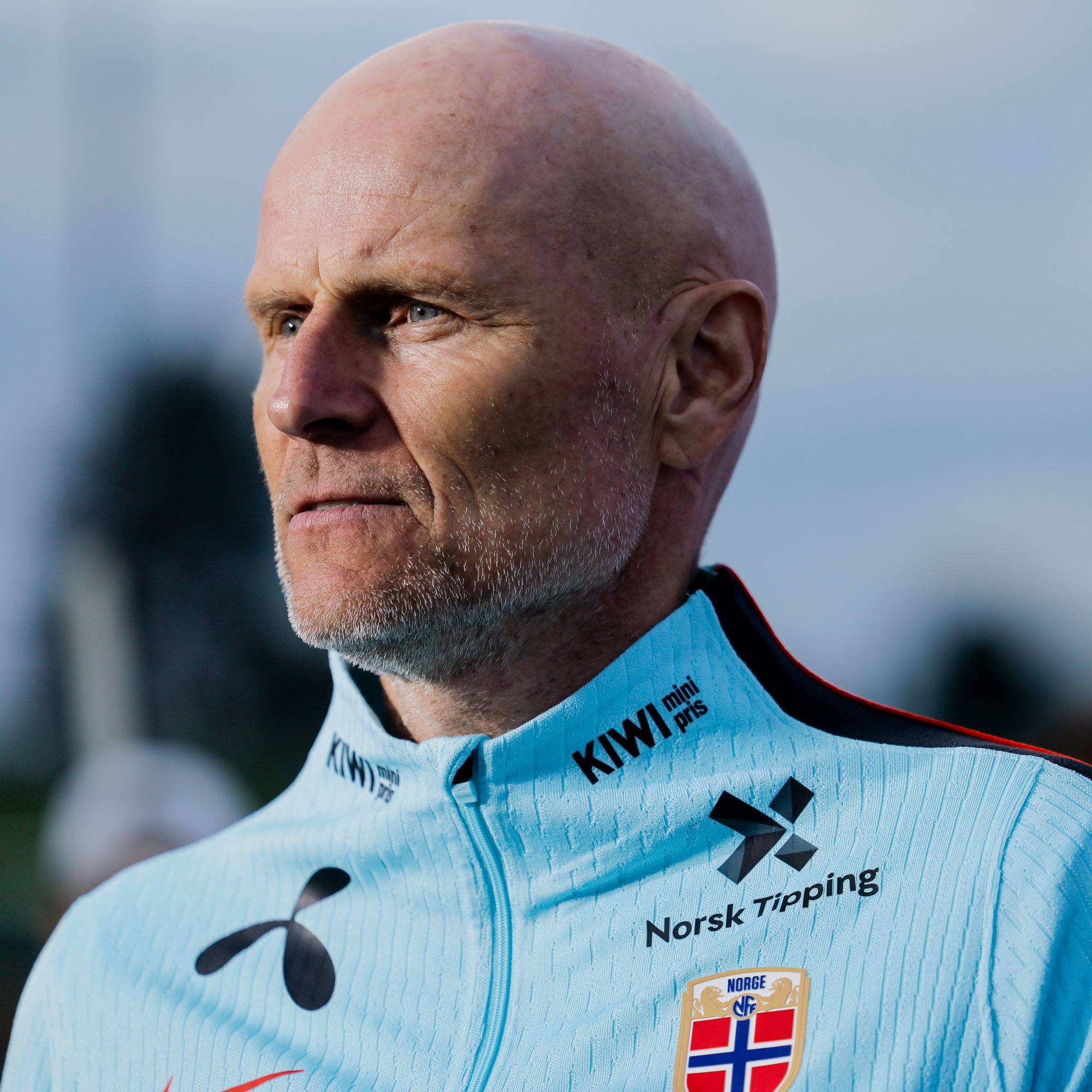 Bald mann i blå treningsjakke med sponsornavn og norsk flagg. Bakgrunnen er utydelig og lys. (Bildebeskrivelsen er laget av en KI-tjeneste)