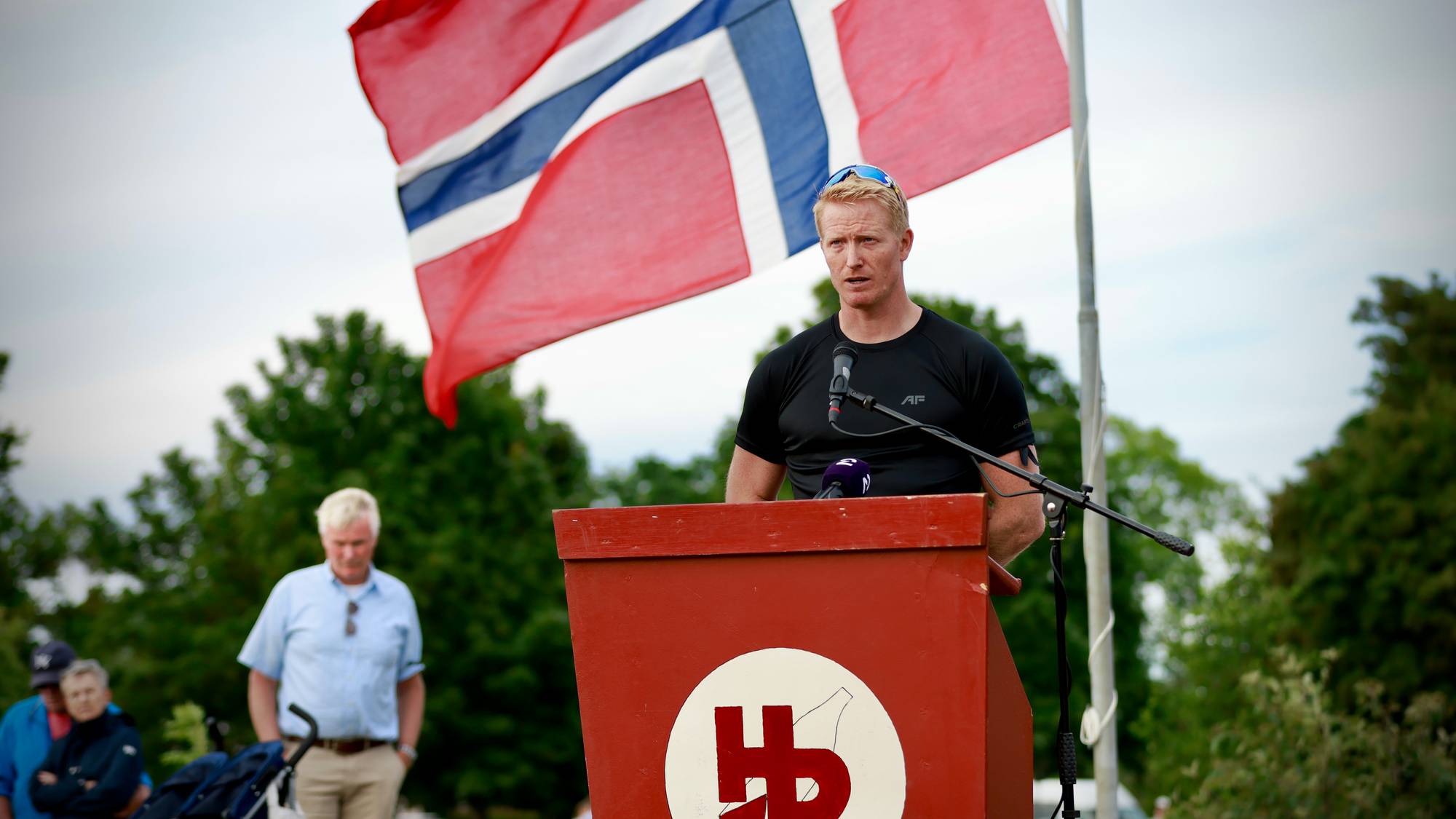 Mann snakker fra en talerstol foran et norsk flagg utendørs.