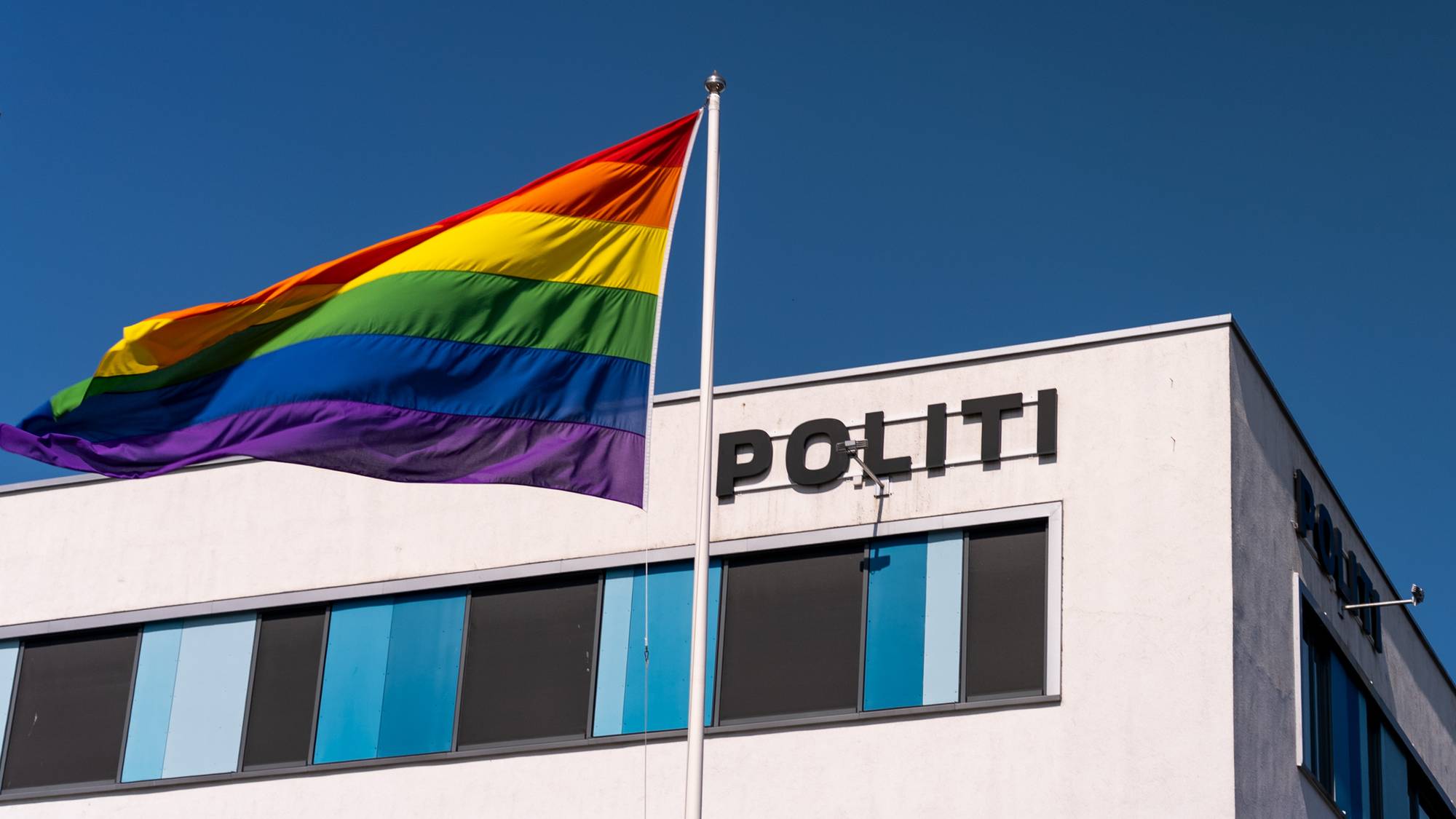 Et regnbueflagg vaier foran en politibygning under en klar, blå himmel.