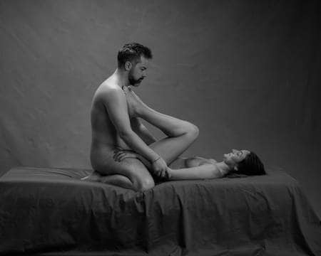 En naken mann med mørkt hår sitter på knærne over en naken dame med langt, mørkt hår som har bena på brystet hans