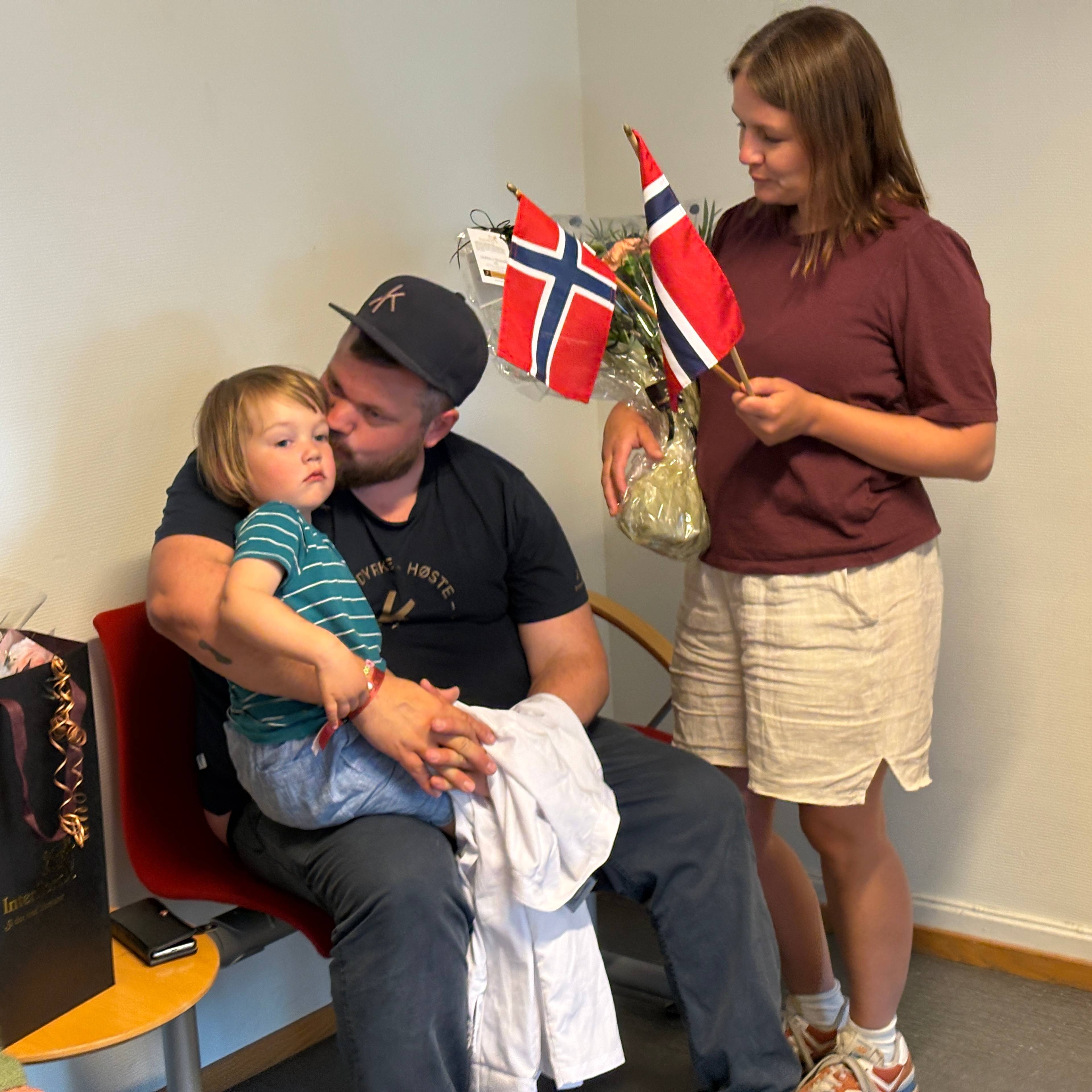 En mann kysser en liten gutt som sitter på fanget hans. En kvinne står ved siden av og holder norske flagg og en gave. Bak dem er det en vegg og en stol. (Bildebeskrivelsen er laget av en KI-tjeneste)