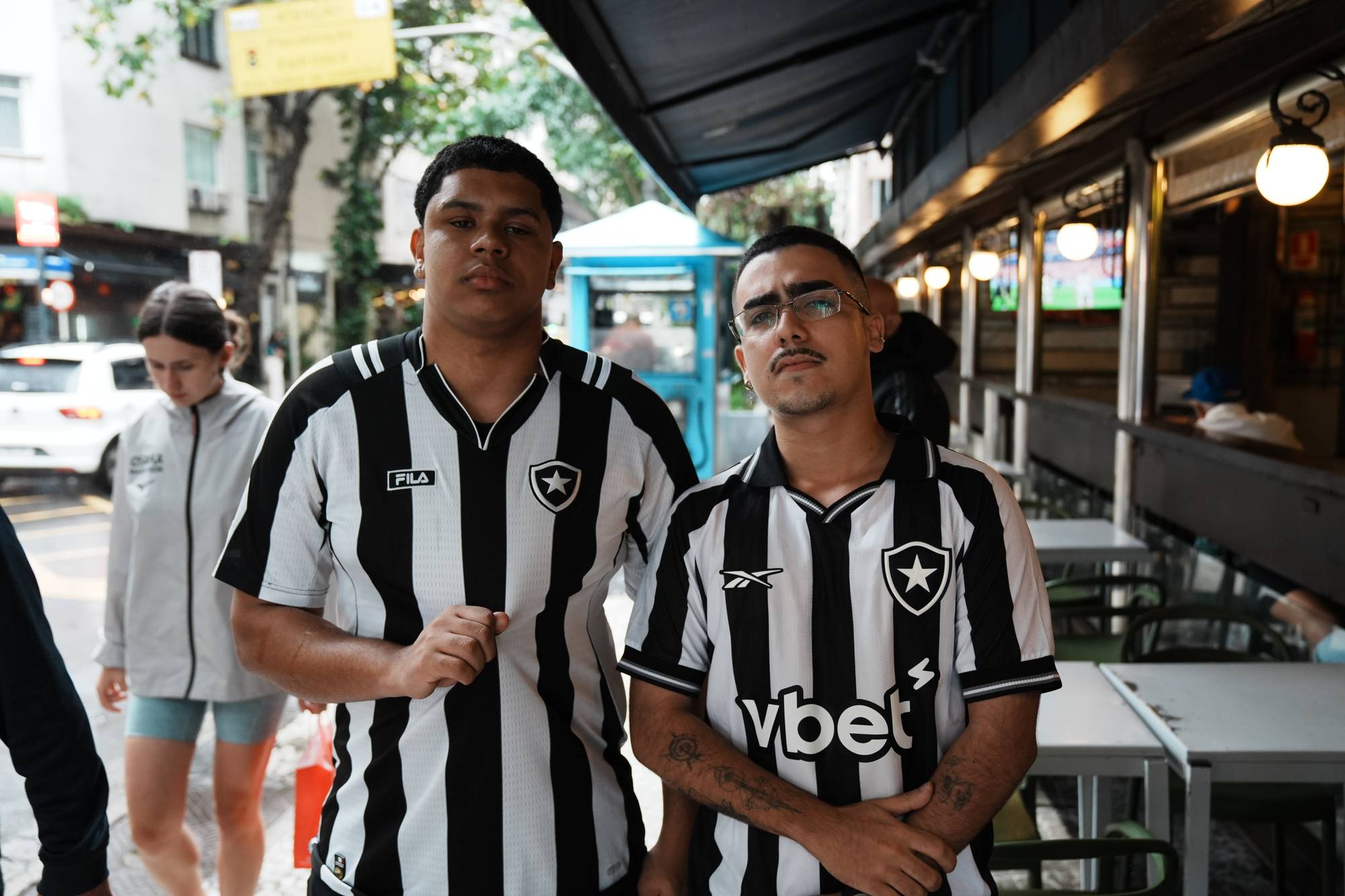 Et portrett av to brasilianske menn i svarte og hvite fotballdrakter som står ved en gate. En kvinne går i bakgrunnen. (Bildebeskrivelsen er laget av en KI-tjeneste)