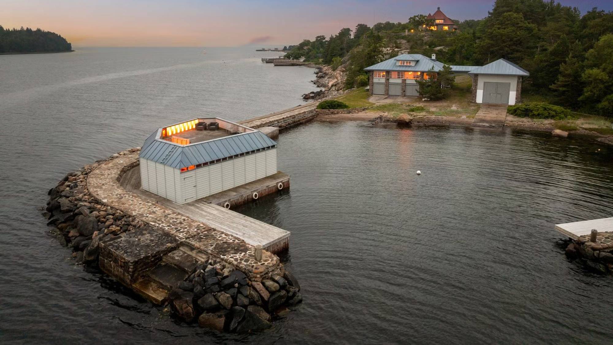 Sjøbod ved en molo og en større bygning ved vannkanten i kystlandskap ved solnedgang. (Bildebeskrivelsen er laget av en KI-tjeneste)