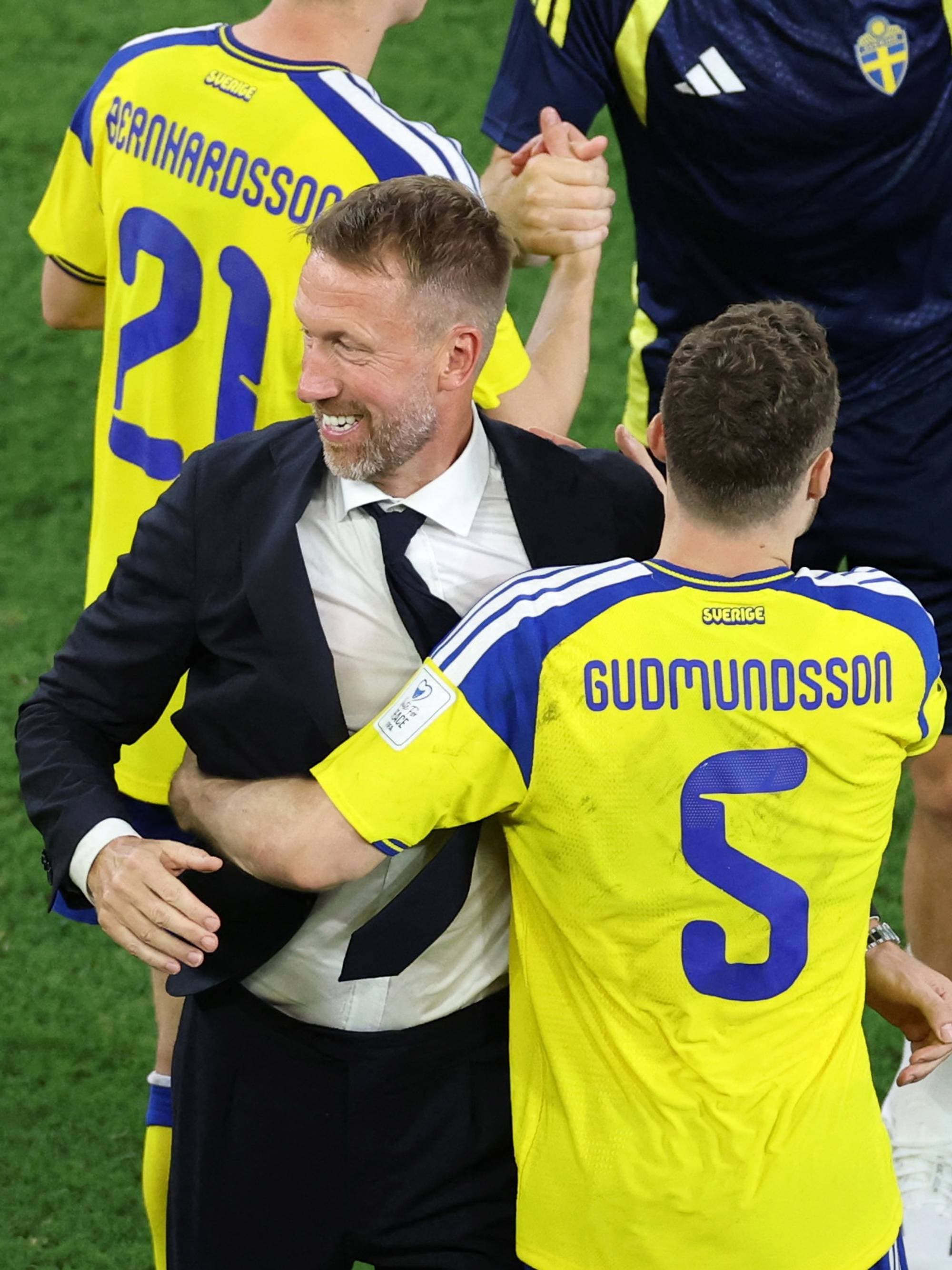 Tre fotballspillere i gule og blå drakter, samt en person i dress, samhandler på en fotballbane. En av spillerne er en landslagstrener. De er på en grønn gressbane. (Bildebeskrivelsen er laget av en KI-tjeneste)