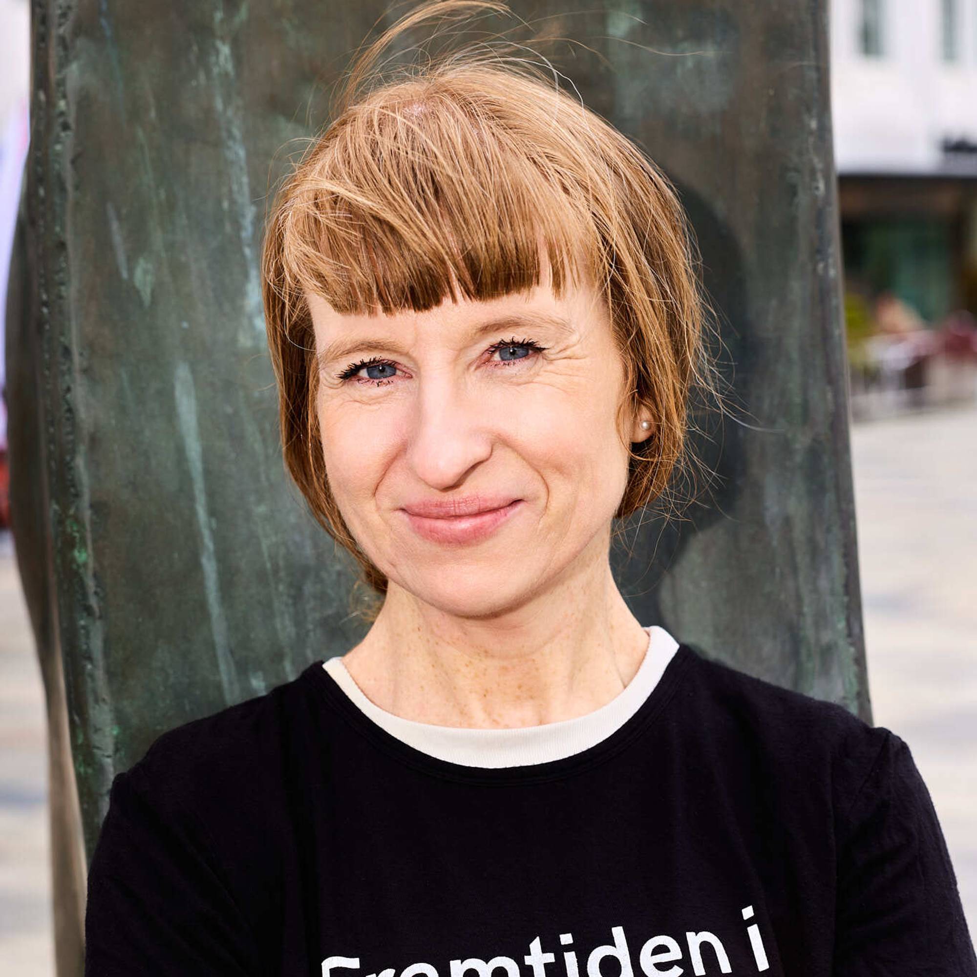 Bildet viser en dame som står med hendene foldet. Hun ghar på seg en t-skjorte med Framtiden i våre hender skreve på brystet. Hun har håret oppsatt og smiler. 