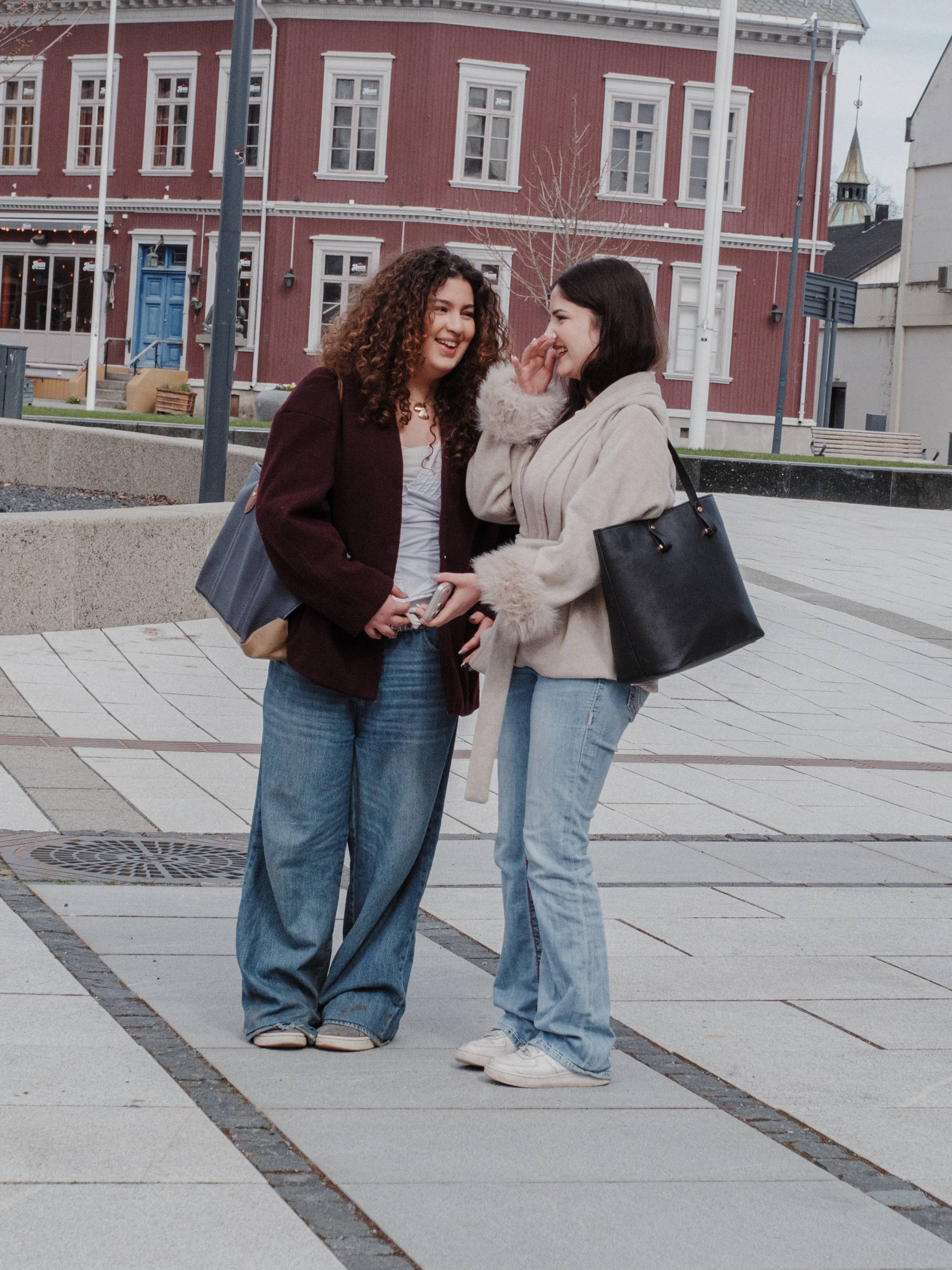 To jenter i jeans, trendy jakker og med vesker på skuldrene ser på hverandre og smiler på en åpen plass i en by.