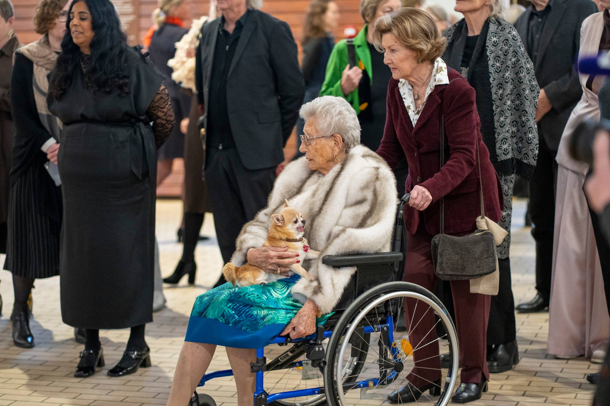 Dronning Sonja triller rullestolen som prinsesse Astrid sitter i, mens hunden Wilma sitter på prinsessens fang.