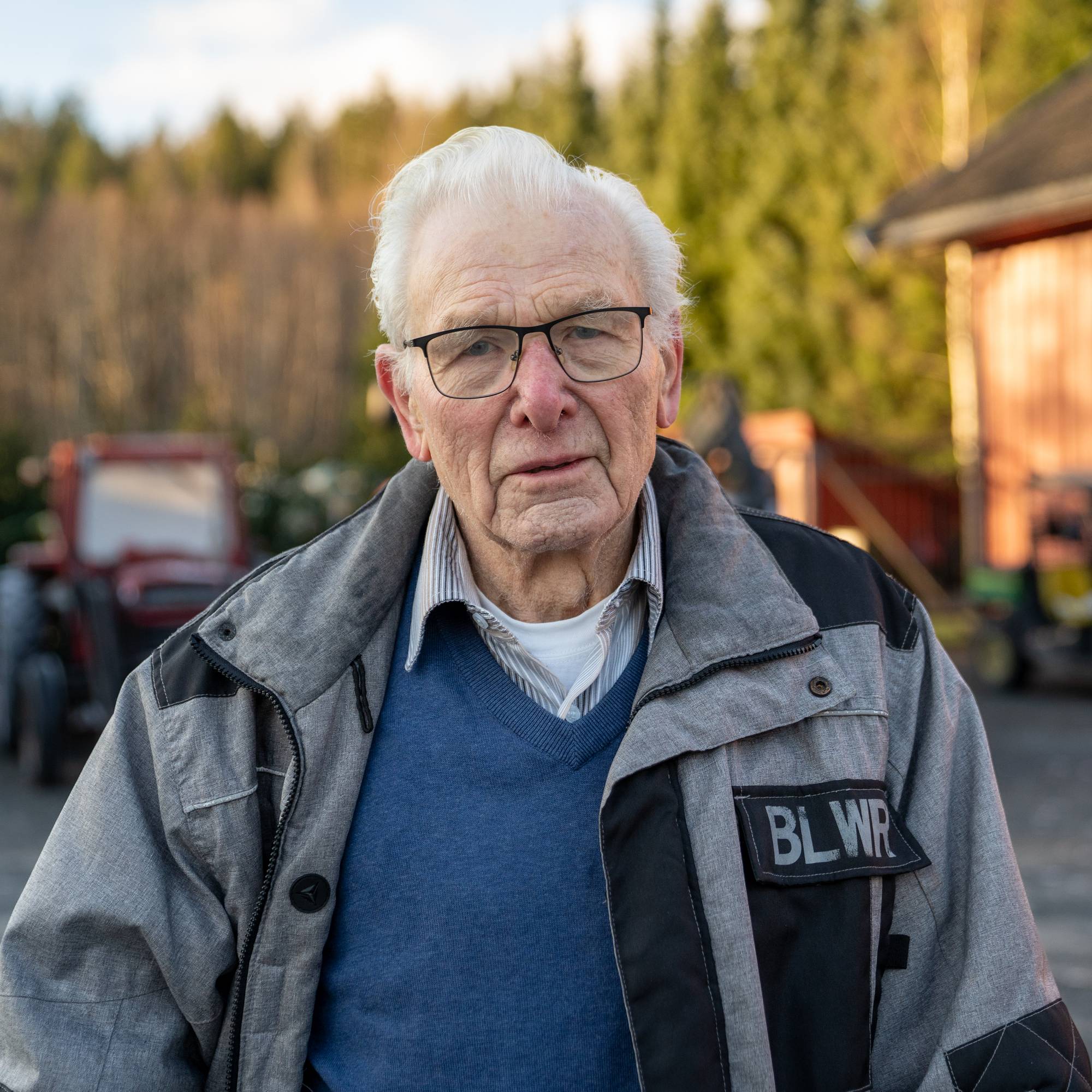 En eldre mann med hvitt hår og grå arbeidsjakke står på en gård. Det er traktorer i bakgrunnen.