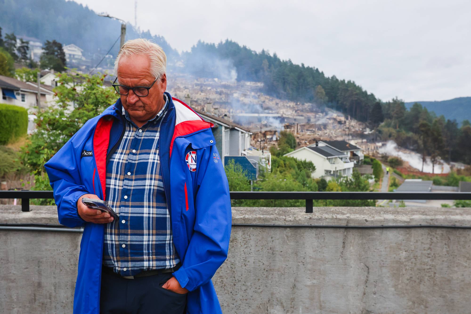 Mann med mobiltelefon foran et boligområde preget av brann. Røyk stiger opp fra ruiner i bakgrunnen. 