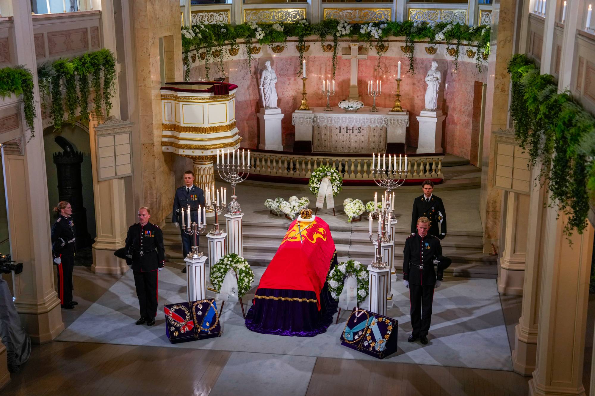Fem kadetter står vakt ved siden av Kong Harald Vs båre. Båren er drapert i kongeflagget og rundt kisten er flere blomsterkranser.