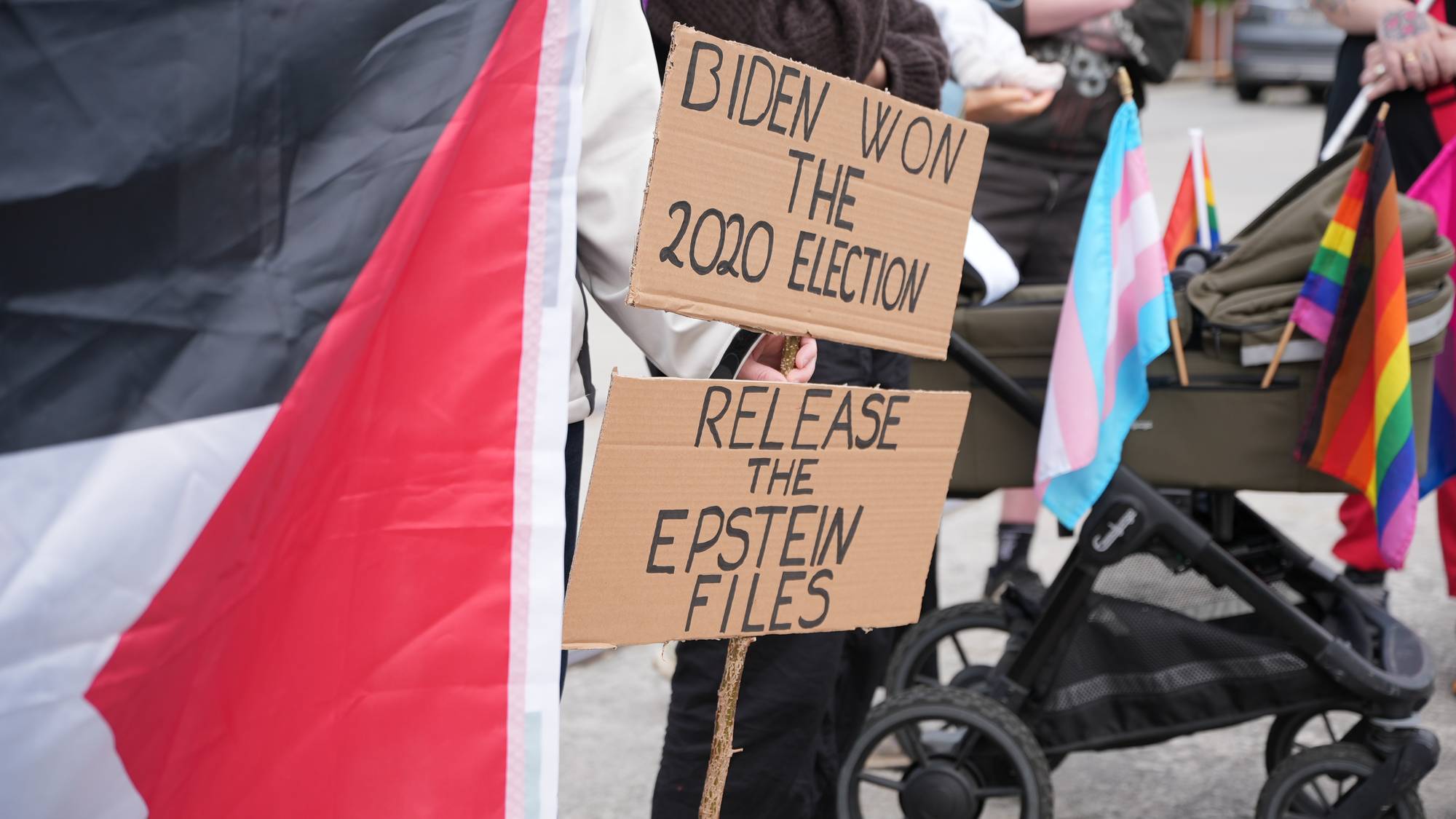 Personer holder plakater med budskapene "BIDEN WON THE 2020 ELECTION" og "RELEASE THE EPSTEIN FILES". En palestinsk flagg er synlig til venstre. I bakgrunnen sees regnbueflagg og et transflagg festet til en barnevogn. Personer er samlet utendørs. (Bildebeskrivelsen er laget av en KI-tjeneste)