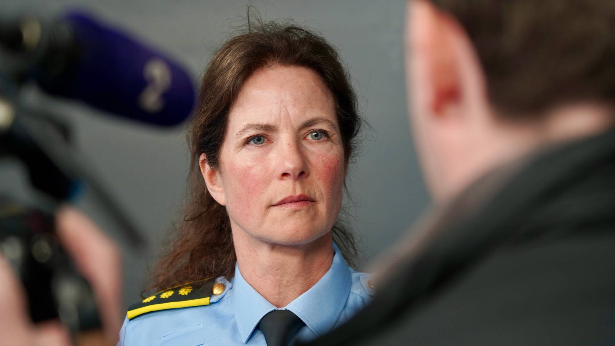 Kvinne i uniform intervjues av journalist.
Bakgrunnen er en grå vegg.
Det er en mikrofon og et kamera i forgrunnen.
Bildet er et fotografi. (Bildebeskrivelsen er laget av en KI-tjeneste)