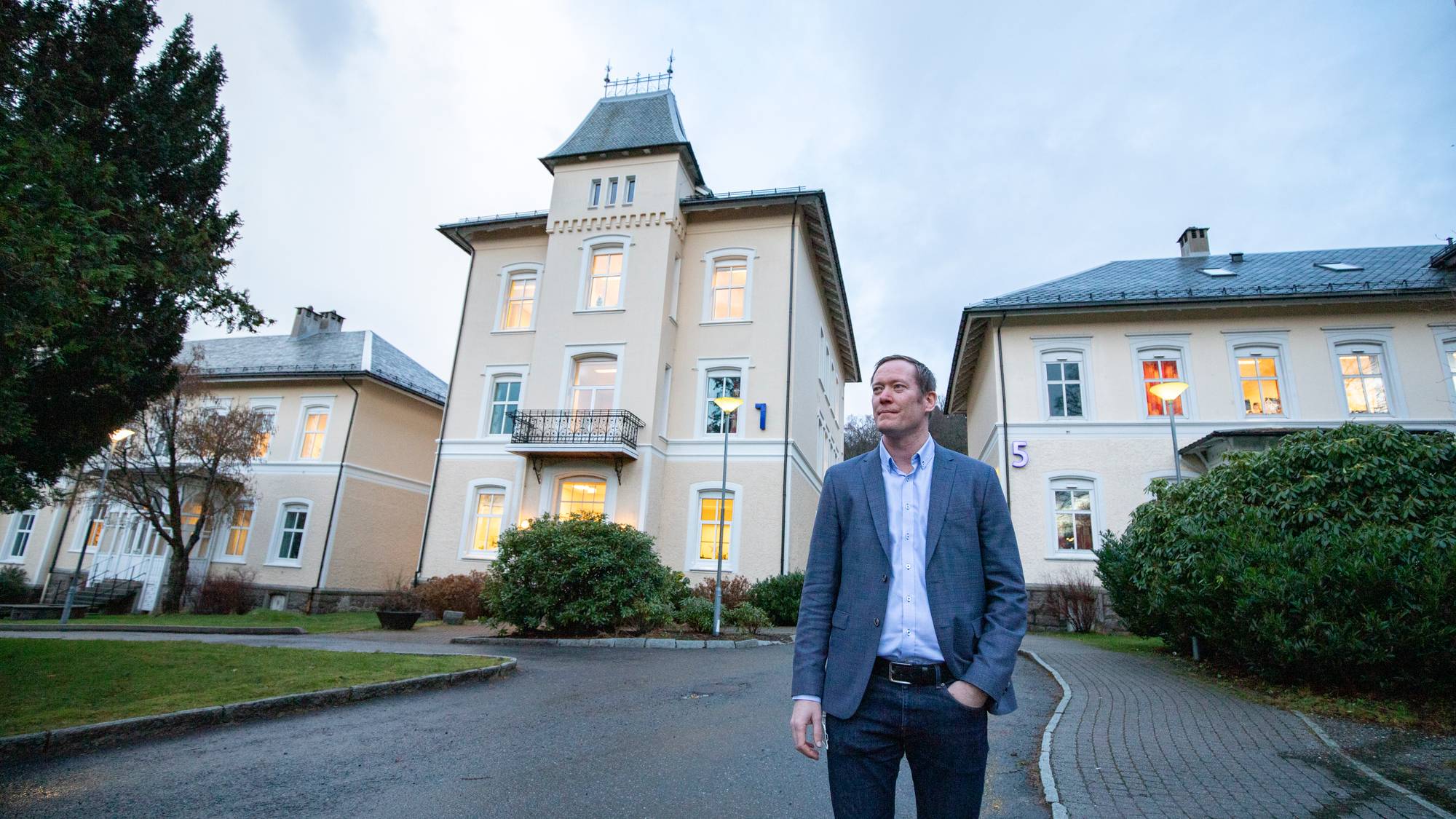 En mann i dressjakke og jeans står utendørs. Han ser til høyre. Bak ham er en lys bygning med flere vinduer og en tårnlignende struktur. Det er trær og busker i forgrunnen og på sidene. 