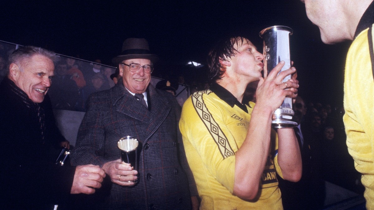 Fotballspiller i gul drakt kysser pokal etter kamp. To menn i dress ser på, en med hatt. (Bildebeskrivelsen er laget av en KI-tjeneste) FOTO: NTB /NTB