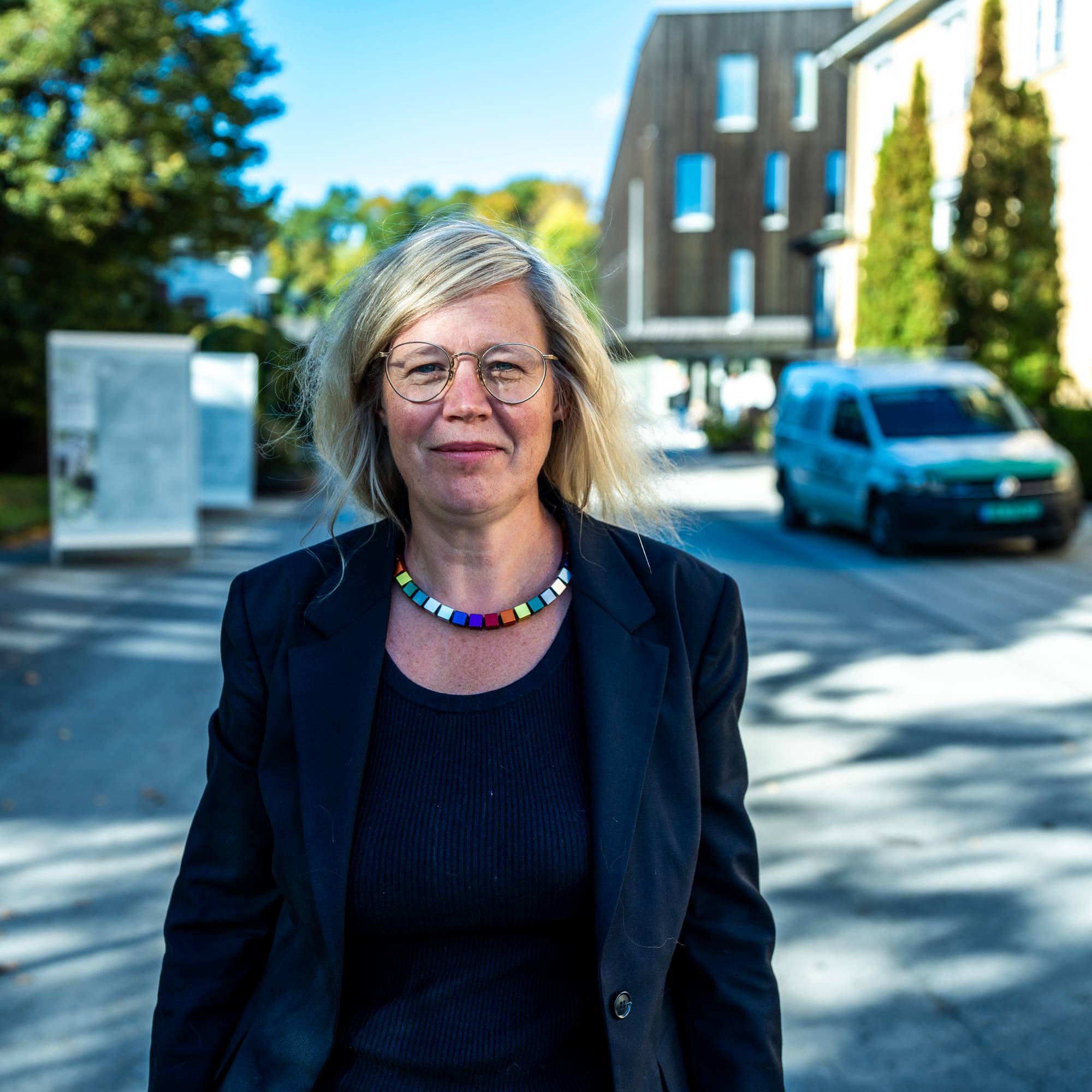 Kvinne med briller og fargerikt smykke står utendørs foran en bygning og en bil. (Bildebeskrivelsen er laget av en KI-tjeneste)