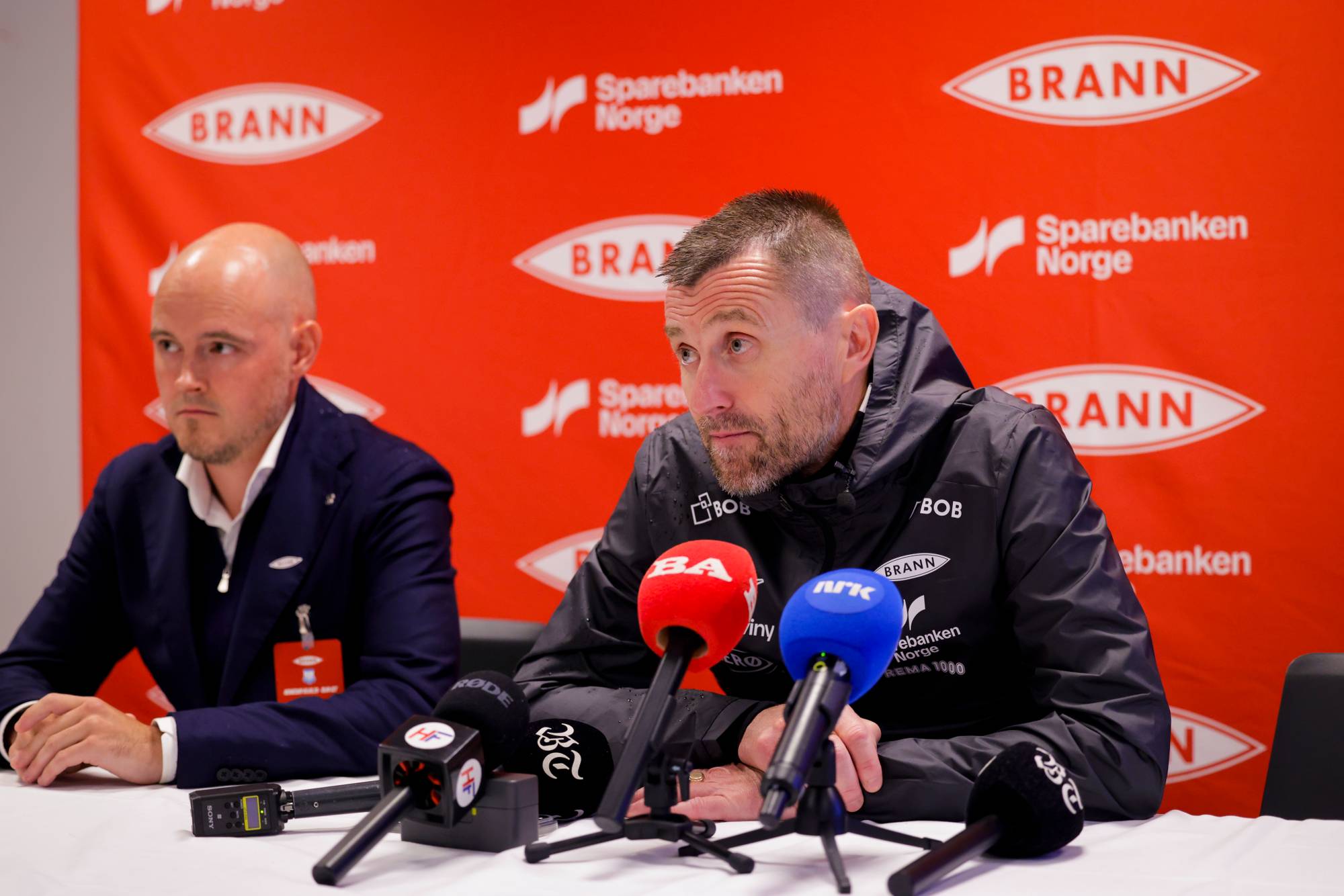 To menn sitter ved et bord med mikrofoner foran seg under en pressekonferanse med Brann-logoer. (Bildebeskrivelsen er laget av en KI-tjeneste)